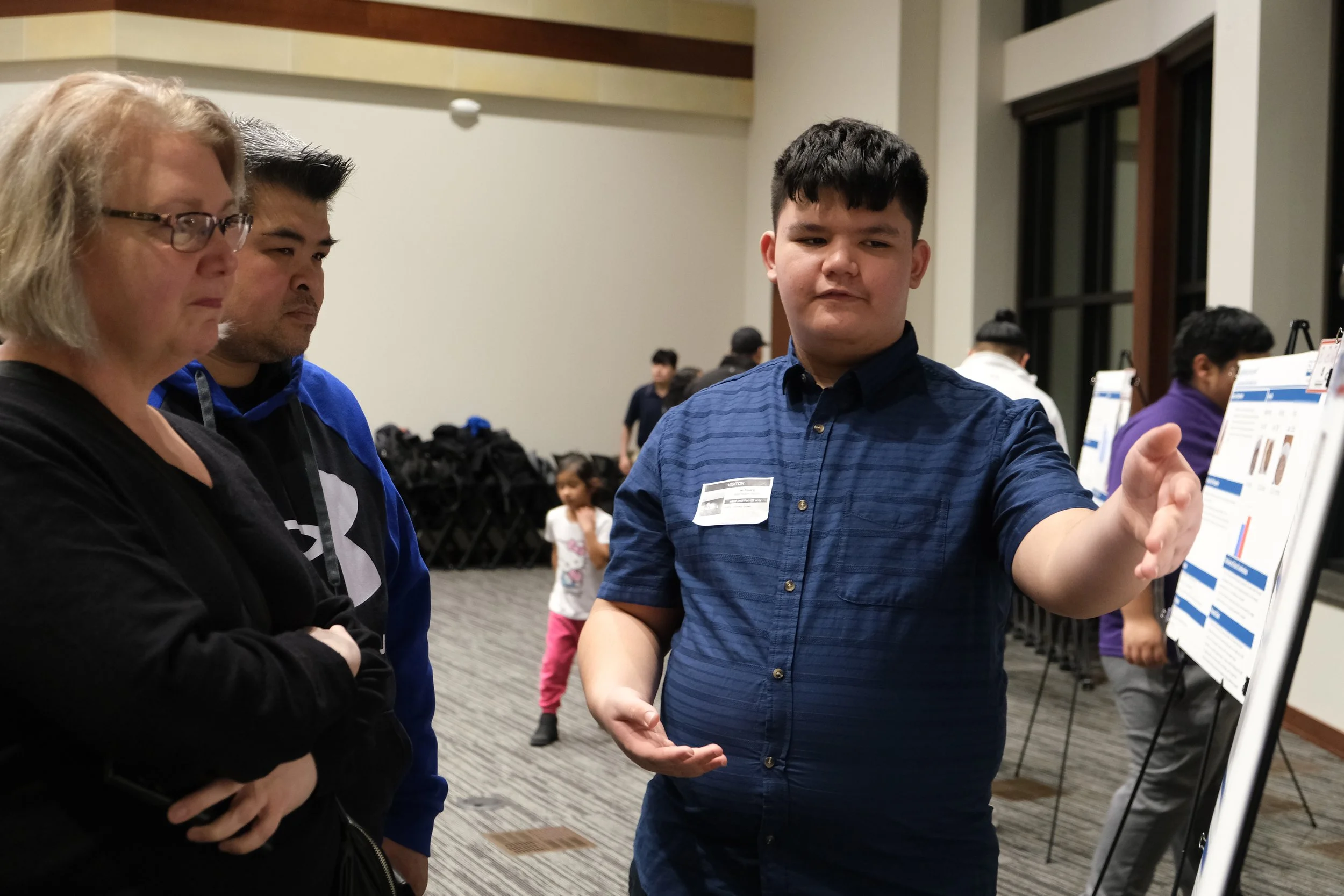 Young man presenting a poster to two adults at an event; people and posters are in the background.