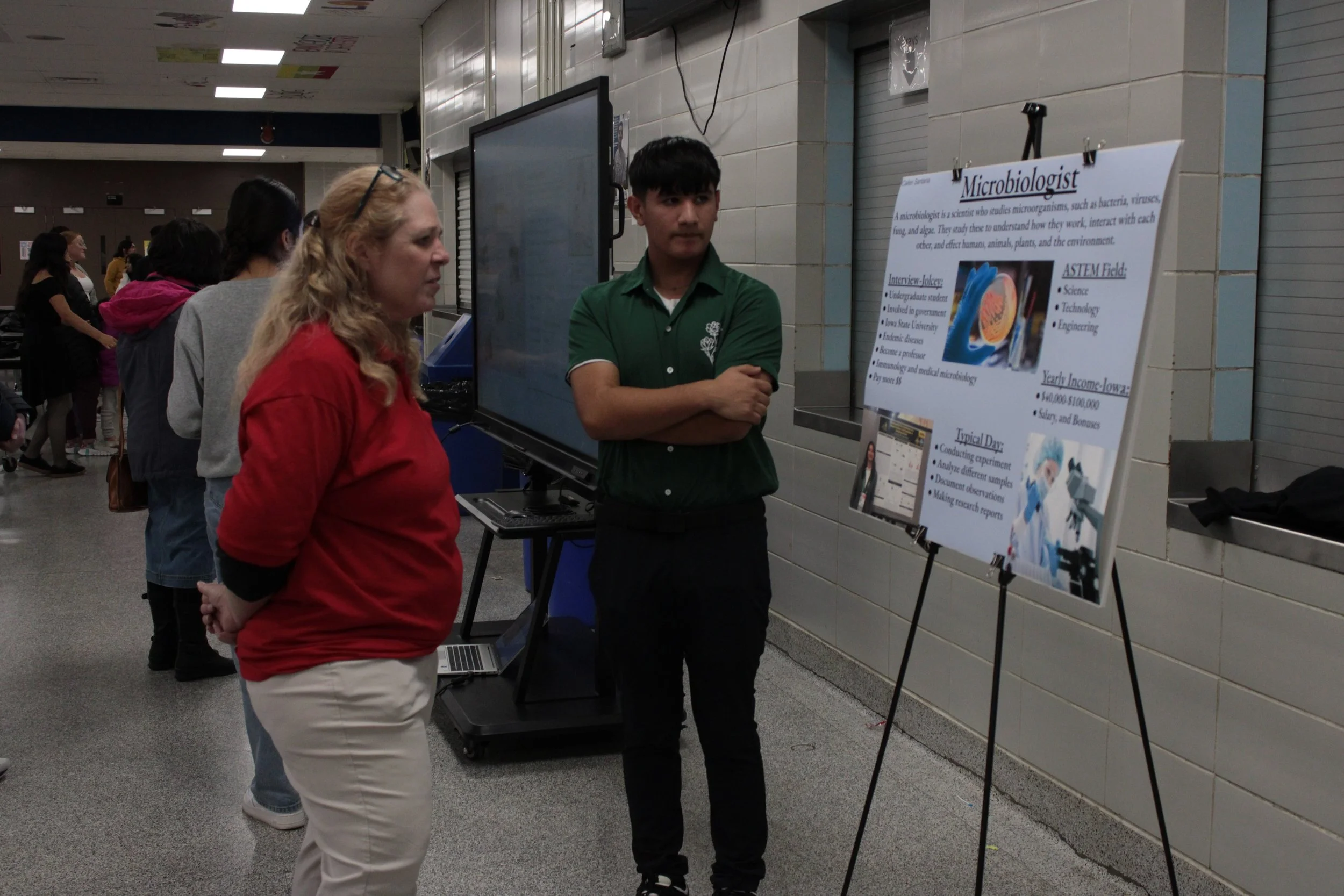 A person in a green shirt presenting a poster about being a microbiologist at an indoor event, with another person in a red shirt listening. The poster includes images and text about the career path, salary, and daily tasks of a microbiologist.