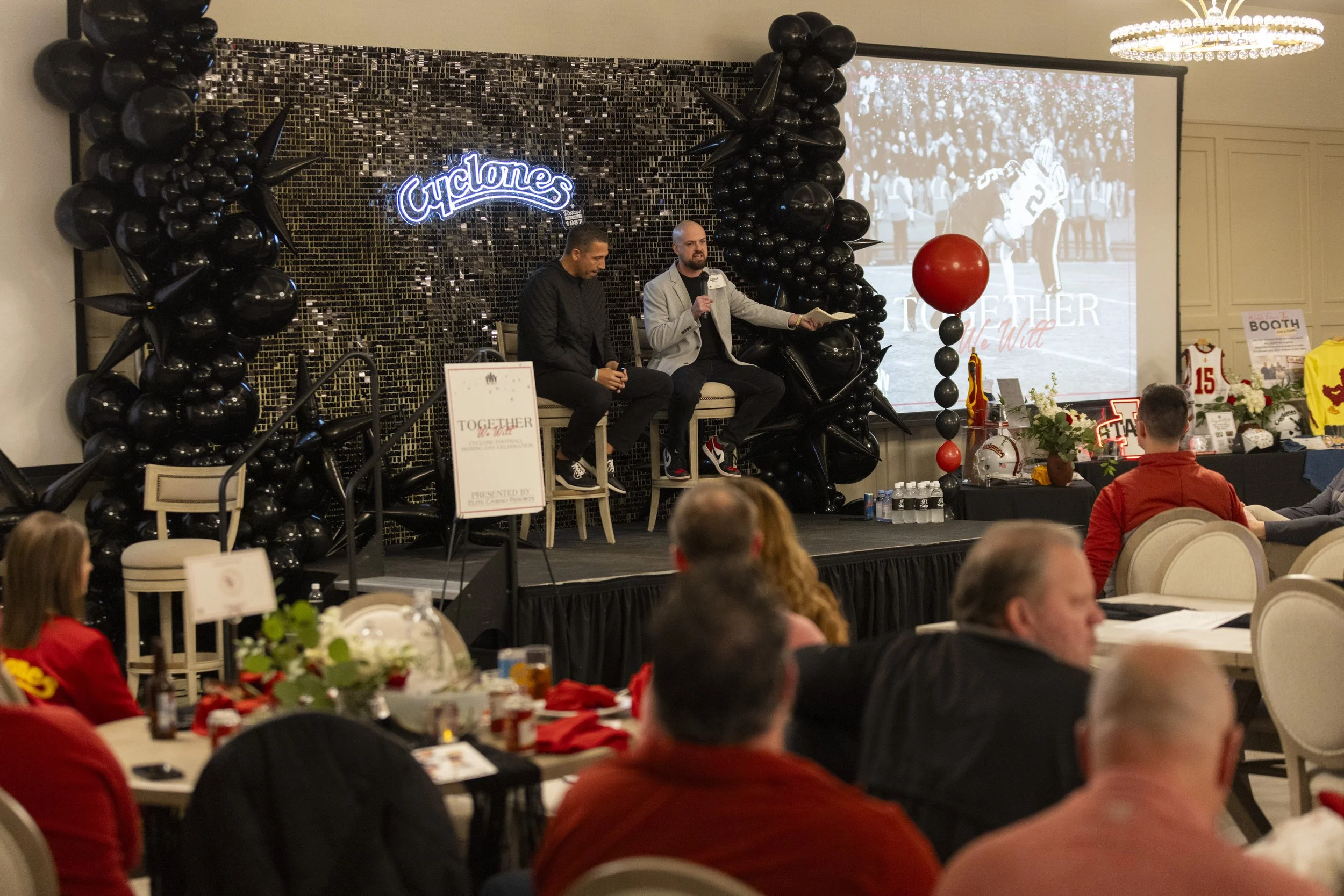 Event with two speakers on stage in front of audience, Cyclones sign, and black balloon decor.