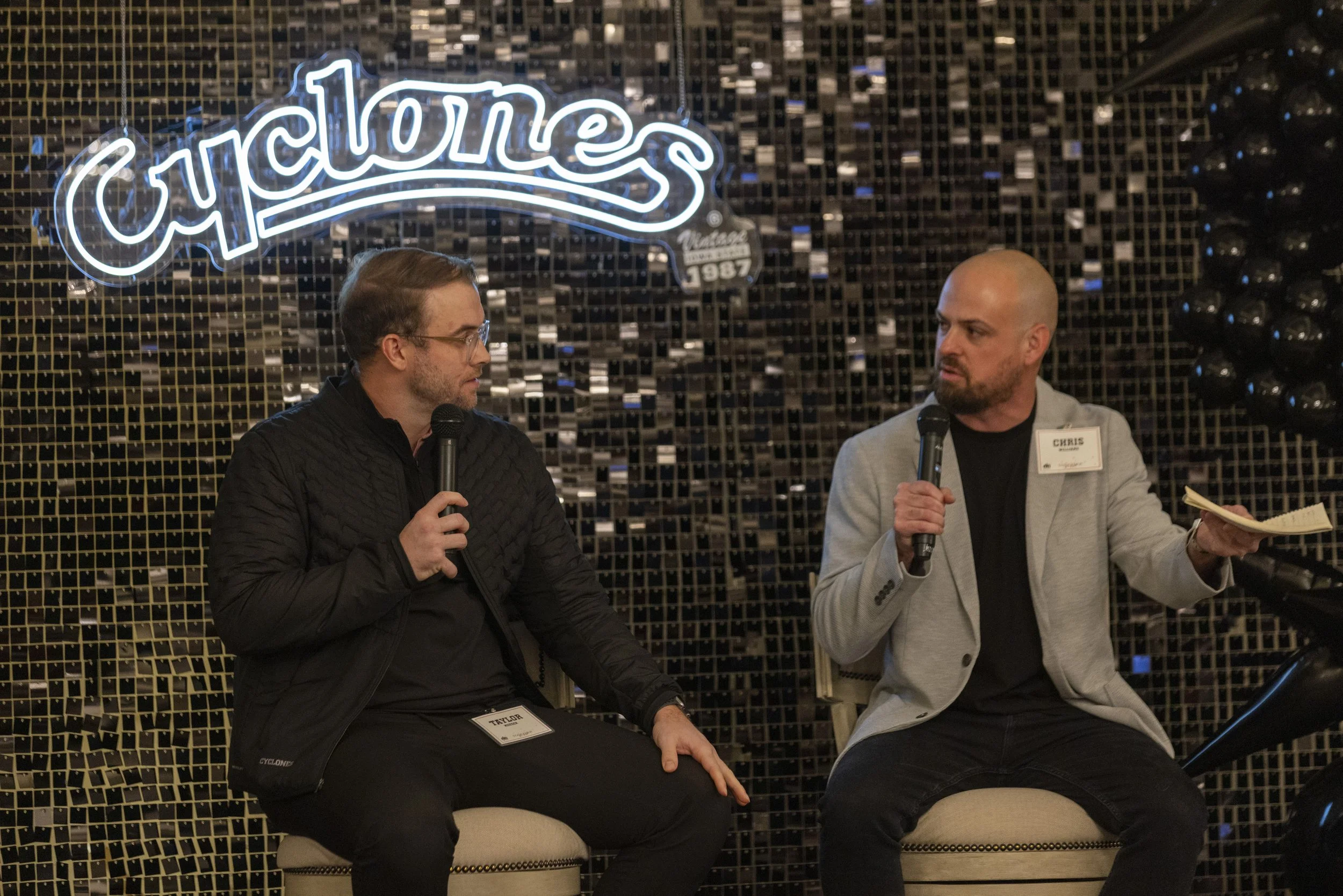 Two men sitting on chairs holding microphones, engaged in conversation. They are in front of a 'Cyclones' sign with a dark, textured background. One man is wearing a dark jacket, and the other is in a light blazer, holding a notepad.