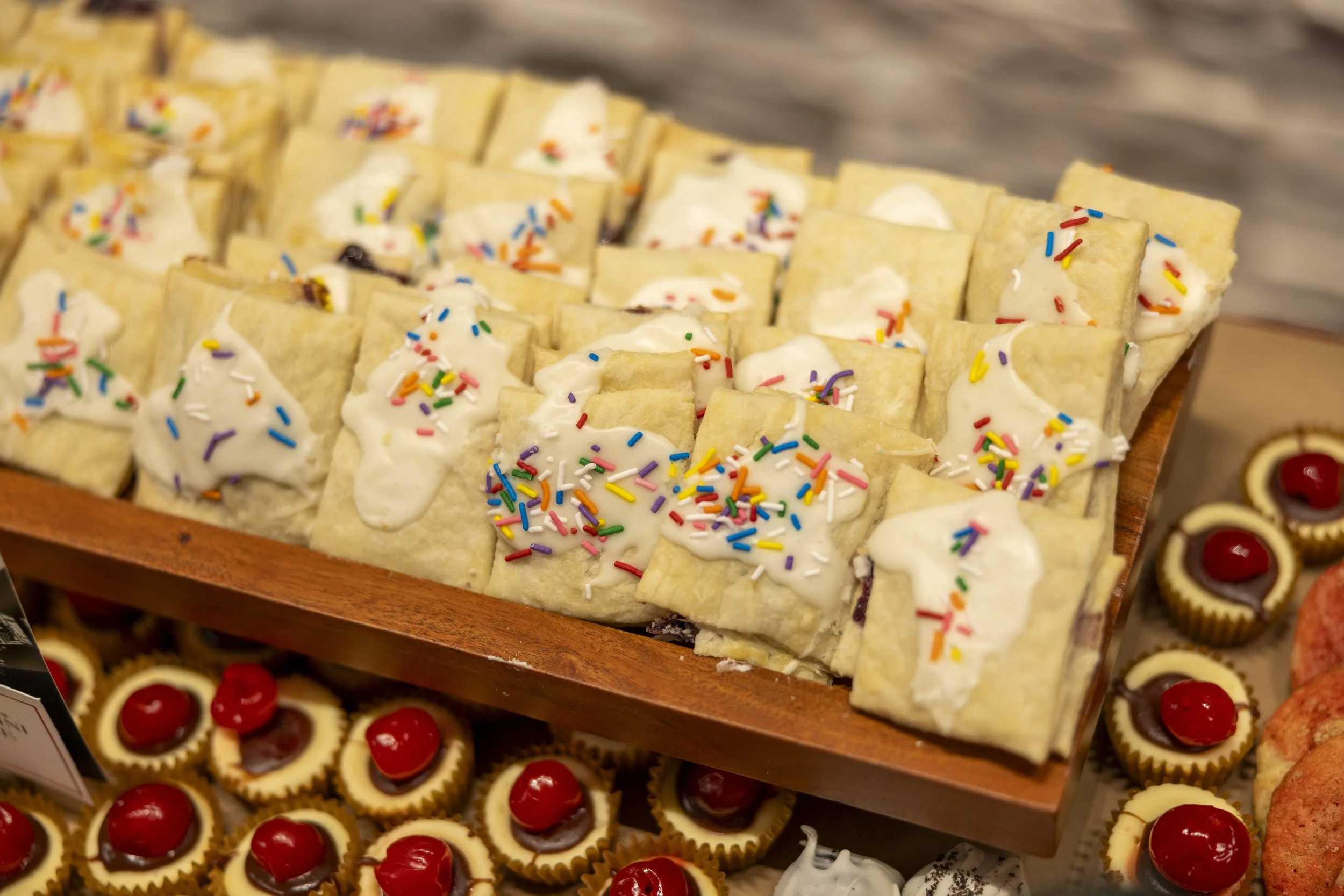 Assorted rectangular pastries with white icing and colorful sprinkles on top, displayed on a wooden tray, surrounded by mini cherry cheesecakes and cookies.