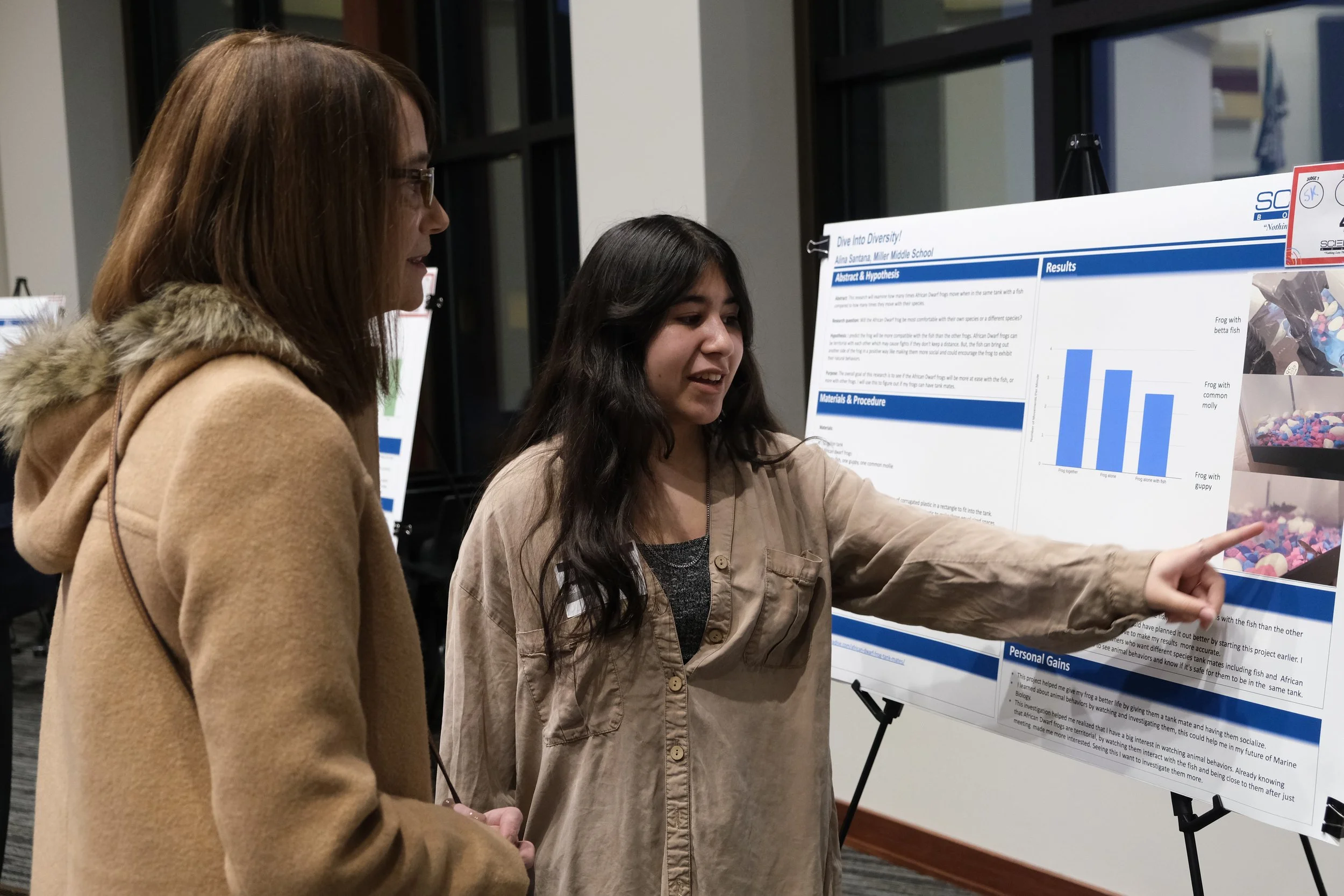 Two people discussing a science fair project displayed on a poster board, featuring charts and images. The person on the right is pointing at the board.