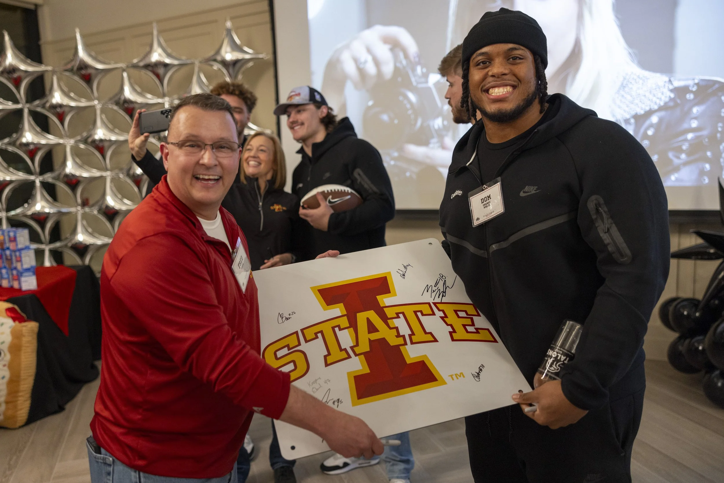 Group of people at an Iowa State event holding a signed poster with the Iowa State logo, smiling, with balloons and screen in the background.