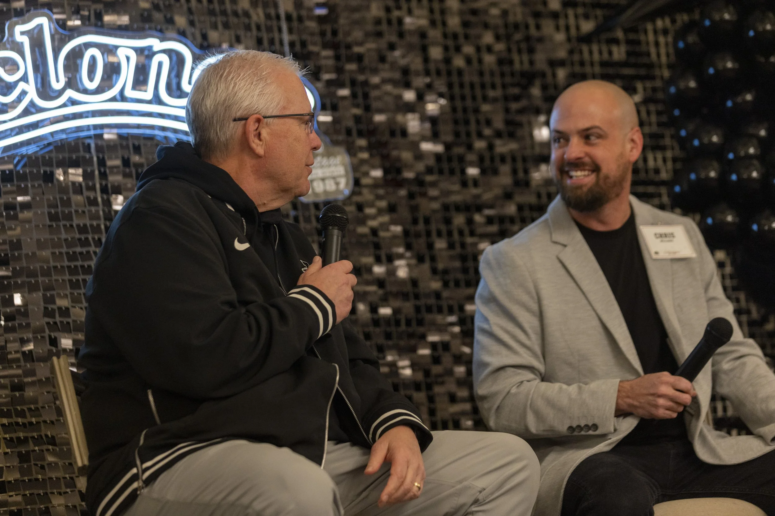 Two men seated, holding microphones, engaging in a conversation with a decorative wall background featuring partial neon text.