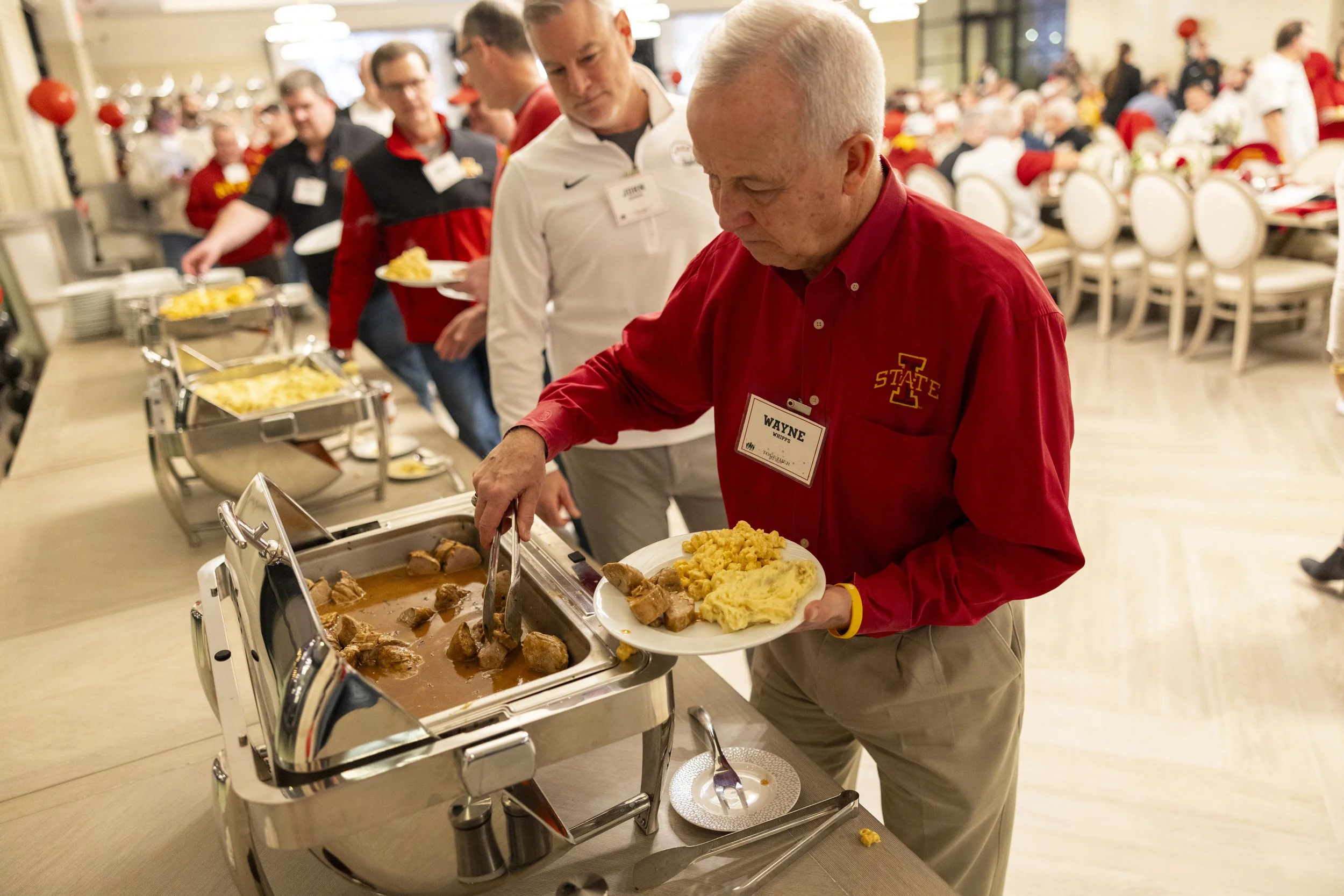 A man in a red shirt serves himself food at a buffet. Others wait in line behind him, holding plates. The setup includes chafing dishes with various foods. The setting appears to be a social or dining event with guests seated in the background.