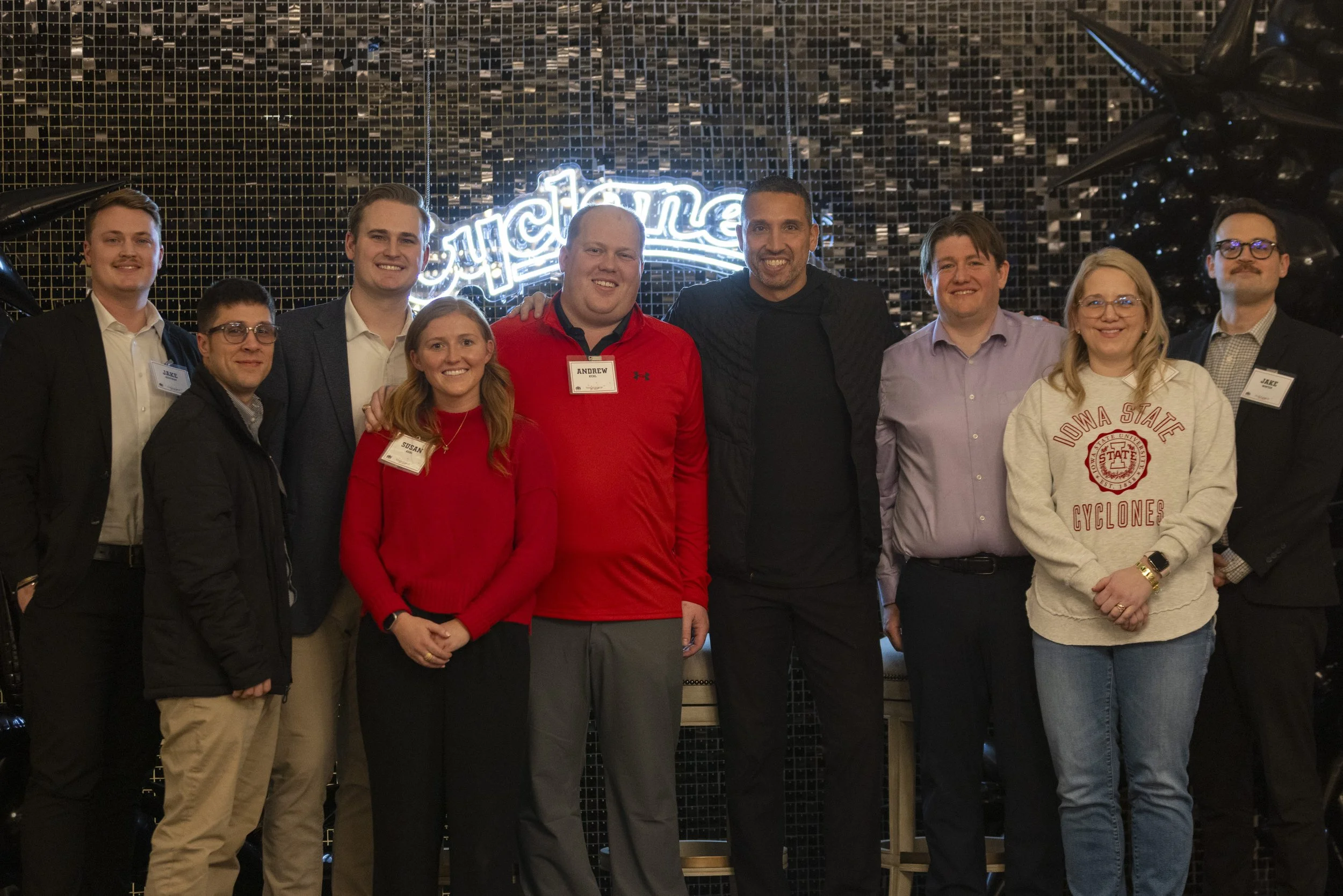 A group of nine people standing in front of a backdrop with "Cyclones" neon sign, some wearing name tags, one person in an Iowa State Cyclones sweatshirt.