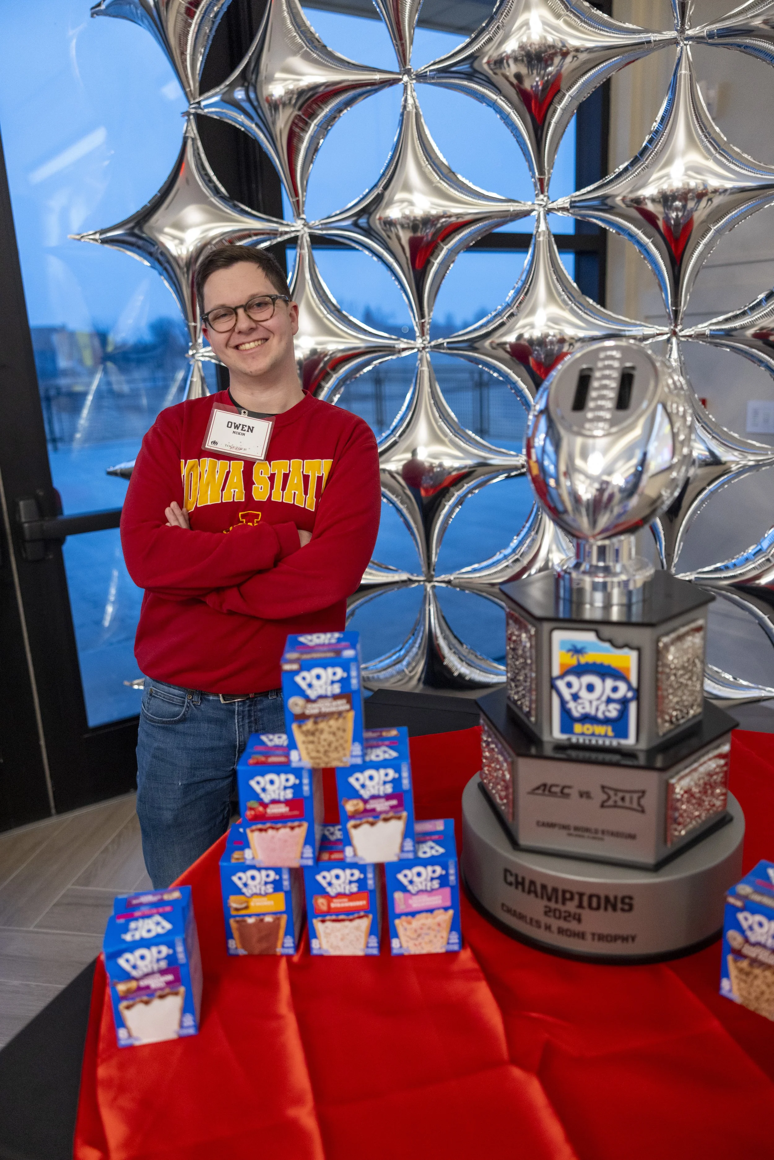 Person wearing an "Iowa State" shirt standing next to a Pop-Tarts display and trophy, with a backdrop of silver star-shaped balloons.