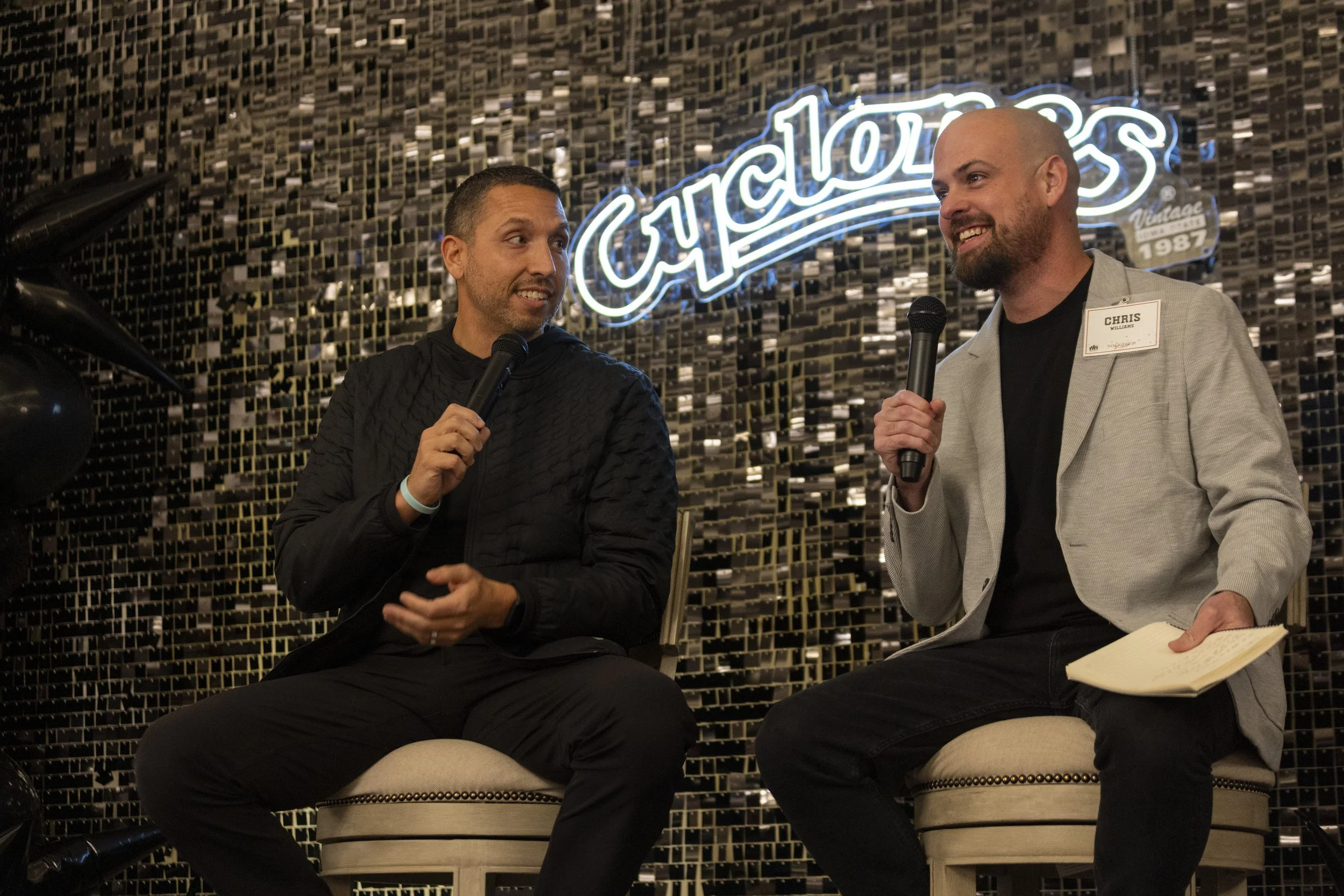Two men sitting and talking on stage, each holding a microphone, in front of a sparkly wall with the word "Cyclones" visible. One man wears a black jacket, the other a gray blazer and a name tag.