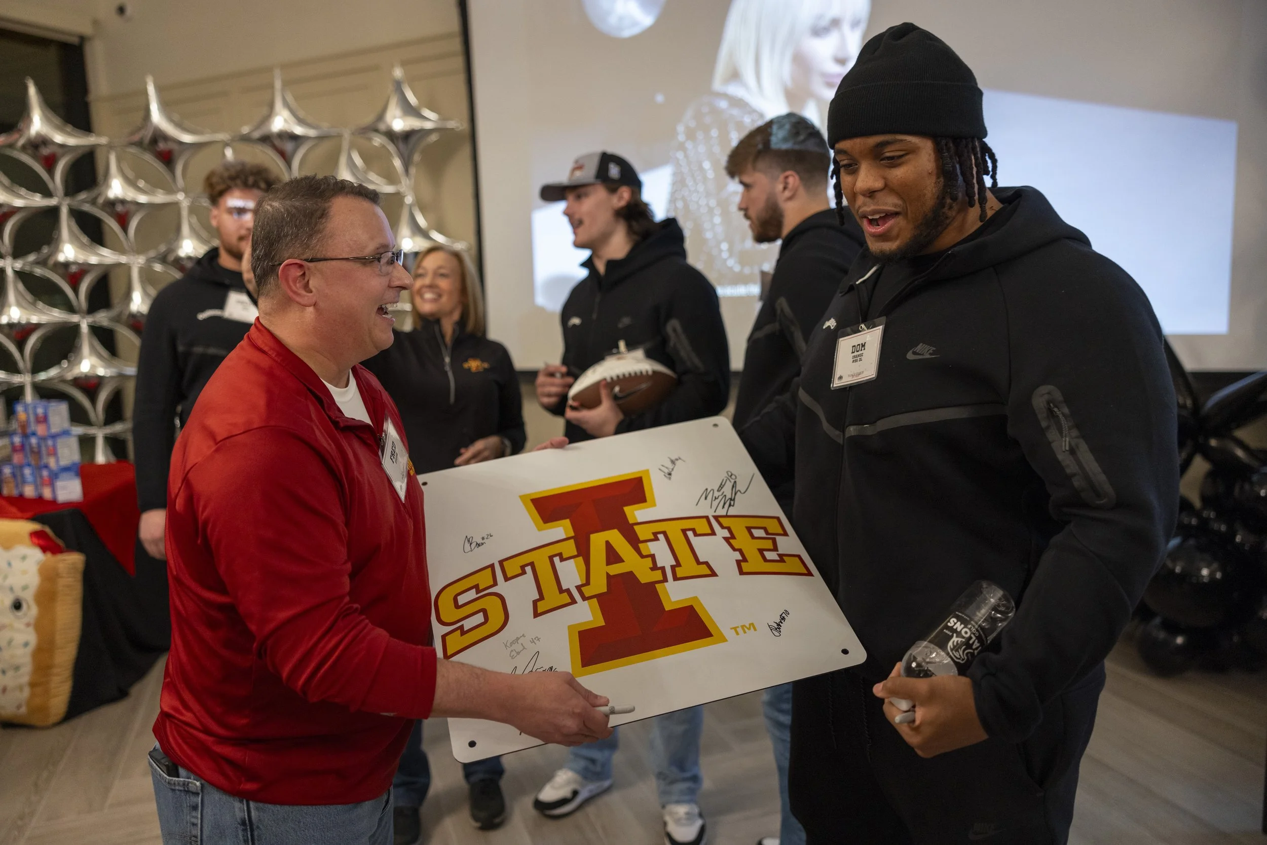 Group of people indoors holding a sign with "I State" logo, football in background, festive decorations, and a woman smiling.