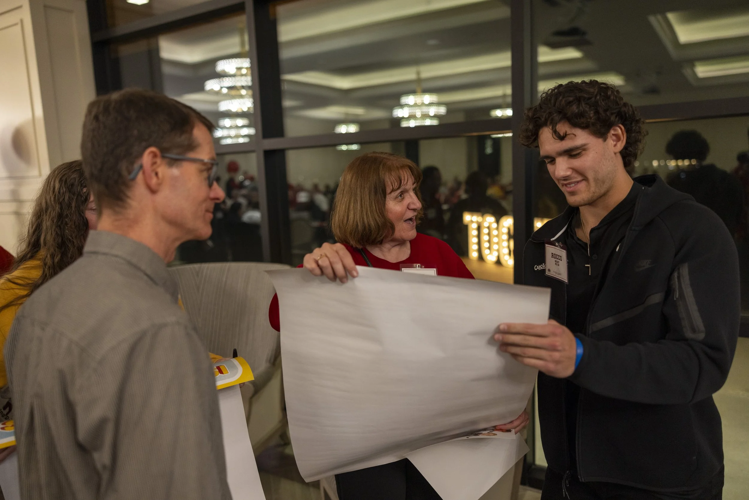 Group of people in a room reviewing a large sheet of paper, with conference name tags and a lit sign in the background.