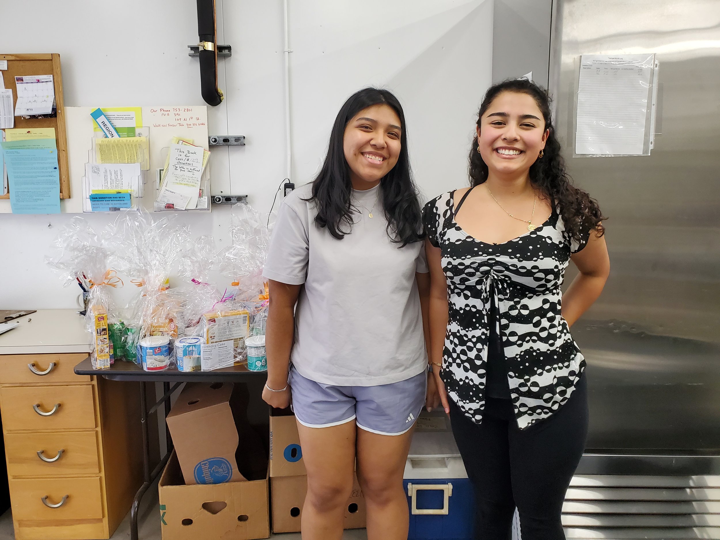 Two smiling women standing in a room with a table full of gift baskets wrapped in plastic. The background includes bulletin boards and metal cupboards. One woman wears a grey shirt and shorts, the other a patterned top and black pants.