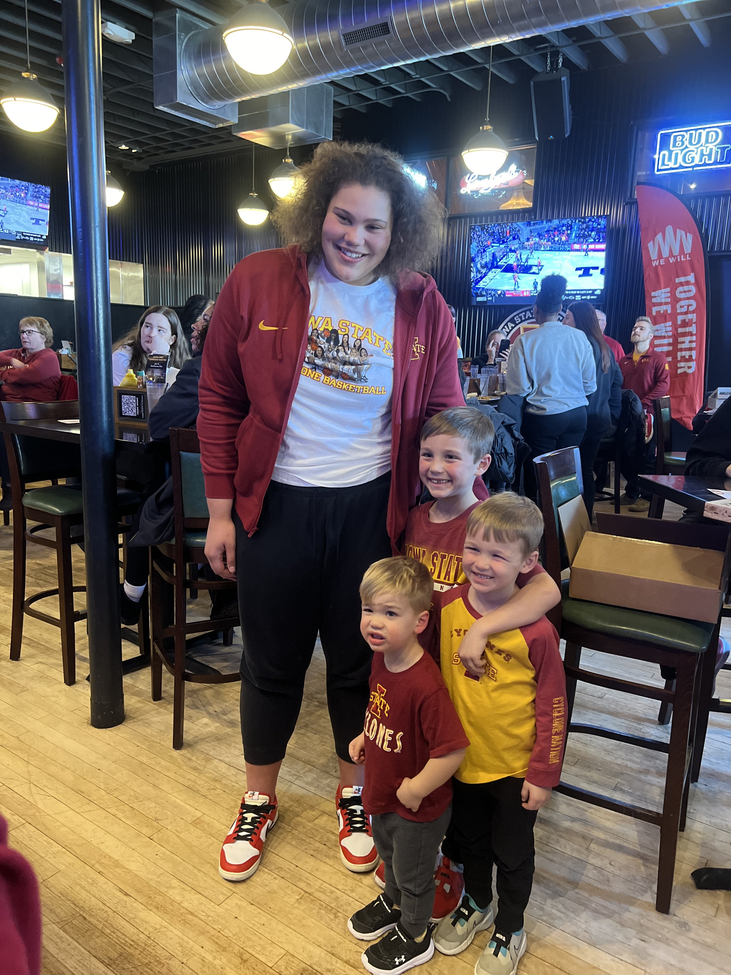 Person wearing Iowa State Cyclones basketball attire standing with three young children in a restaurant with sports decor.