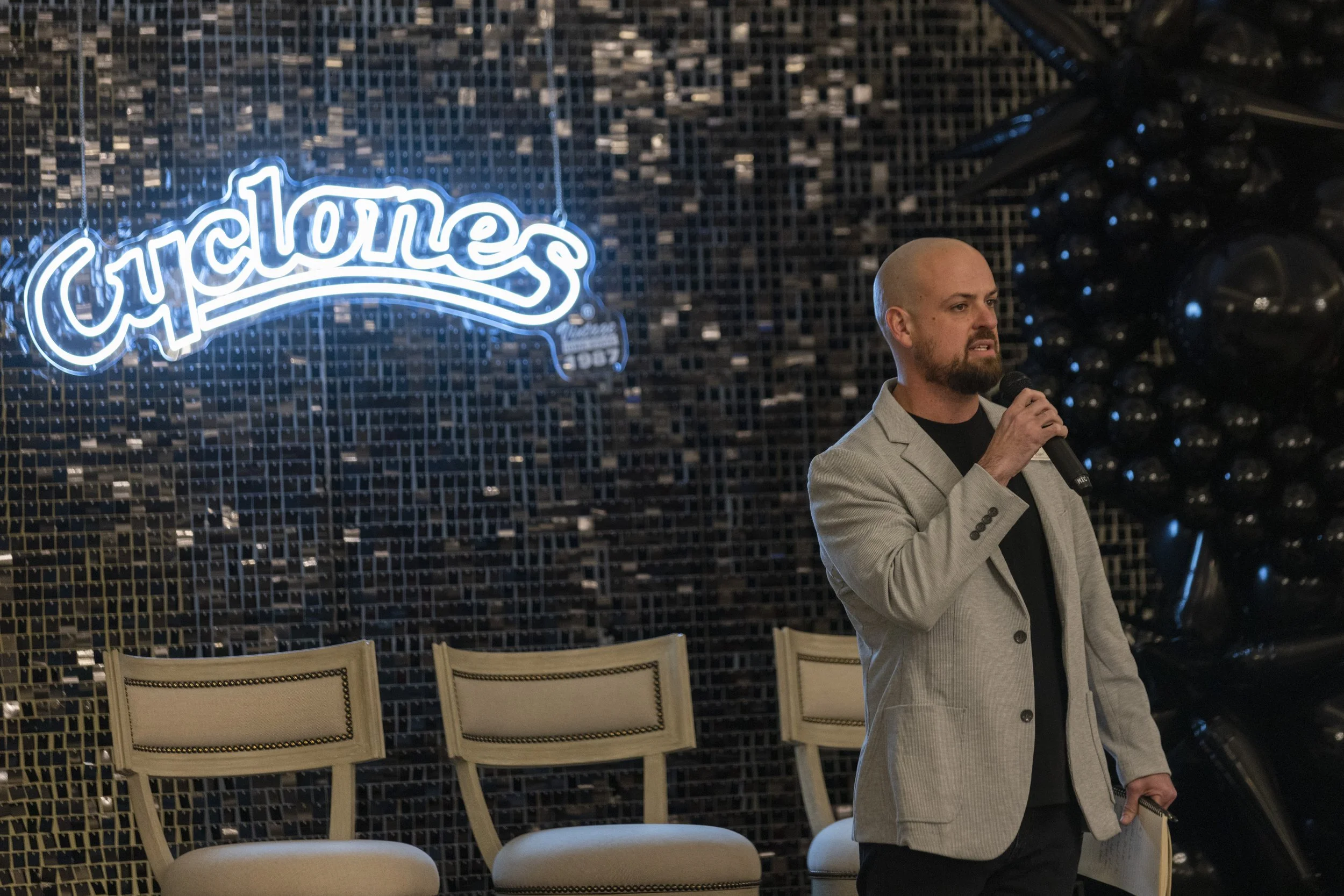 Person speaking into a microphone in front of a "Cyclones" neon sign and empty chairs.