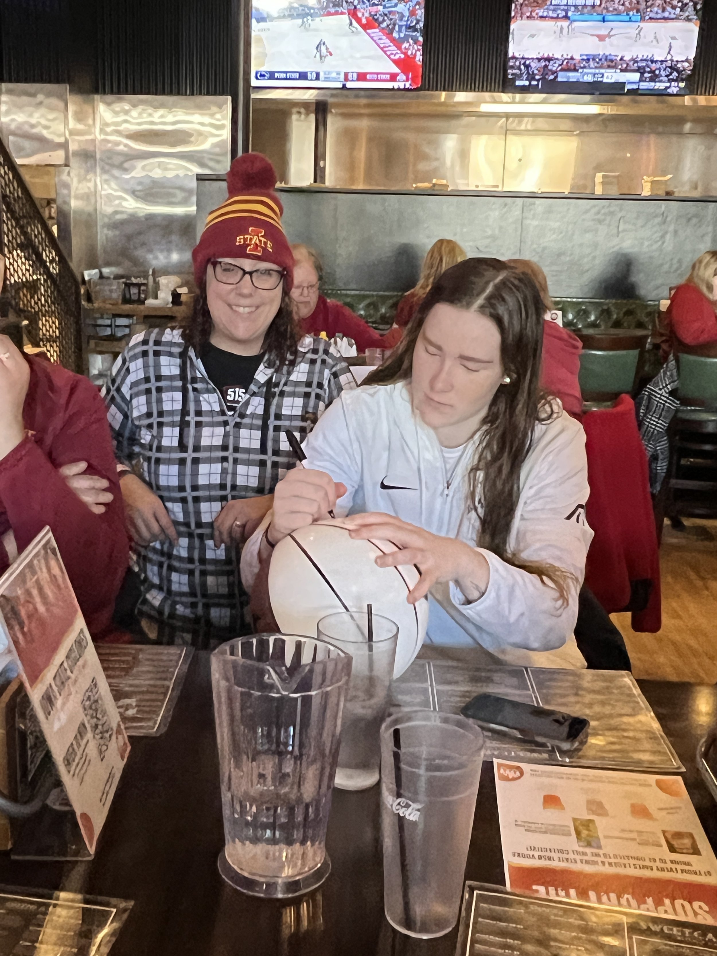Person signing a basketball while another person watches in a sports bar with TVs showing basketball games.