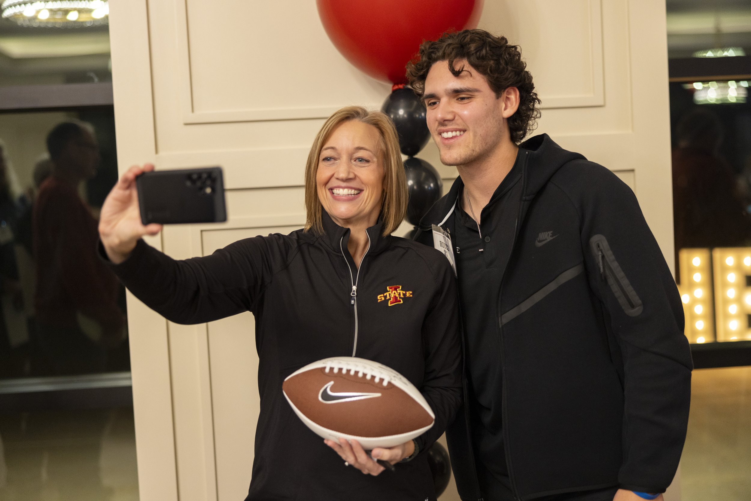 Two people taking a selfie; the one on the left holds a football and wears a jacket with a "State" logo. They are smiling and standing in front of red and black balloons.