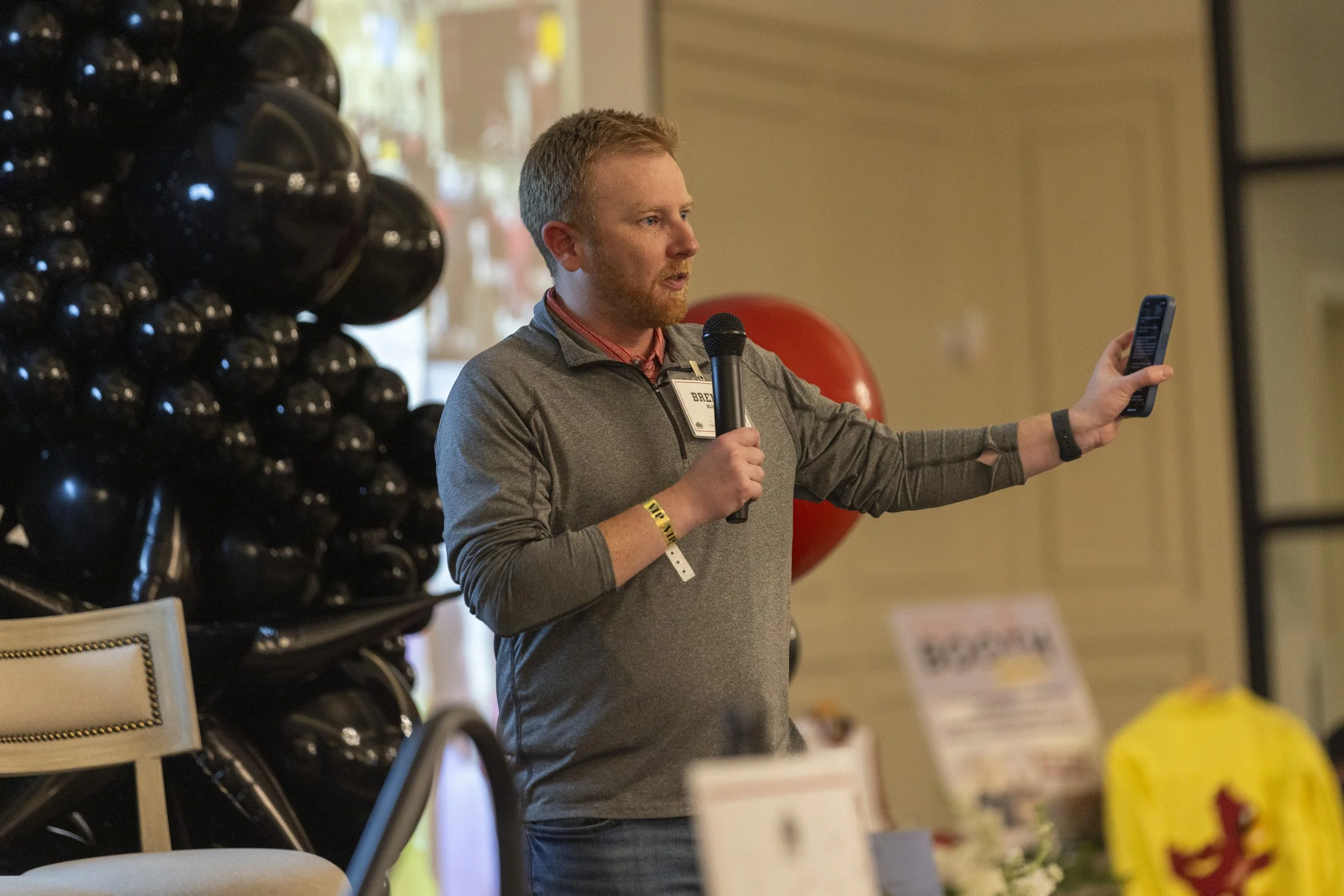 Man speaking into a microphone while holding a smartphone, with black balloons and a presentation screen in the background.