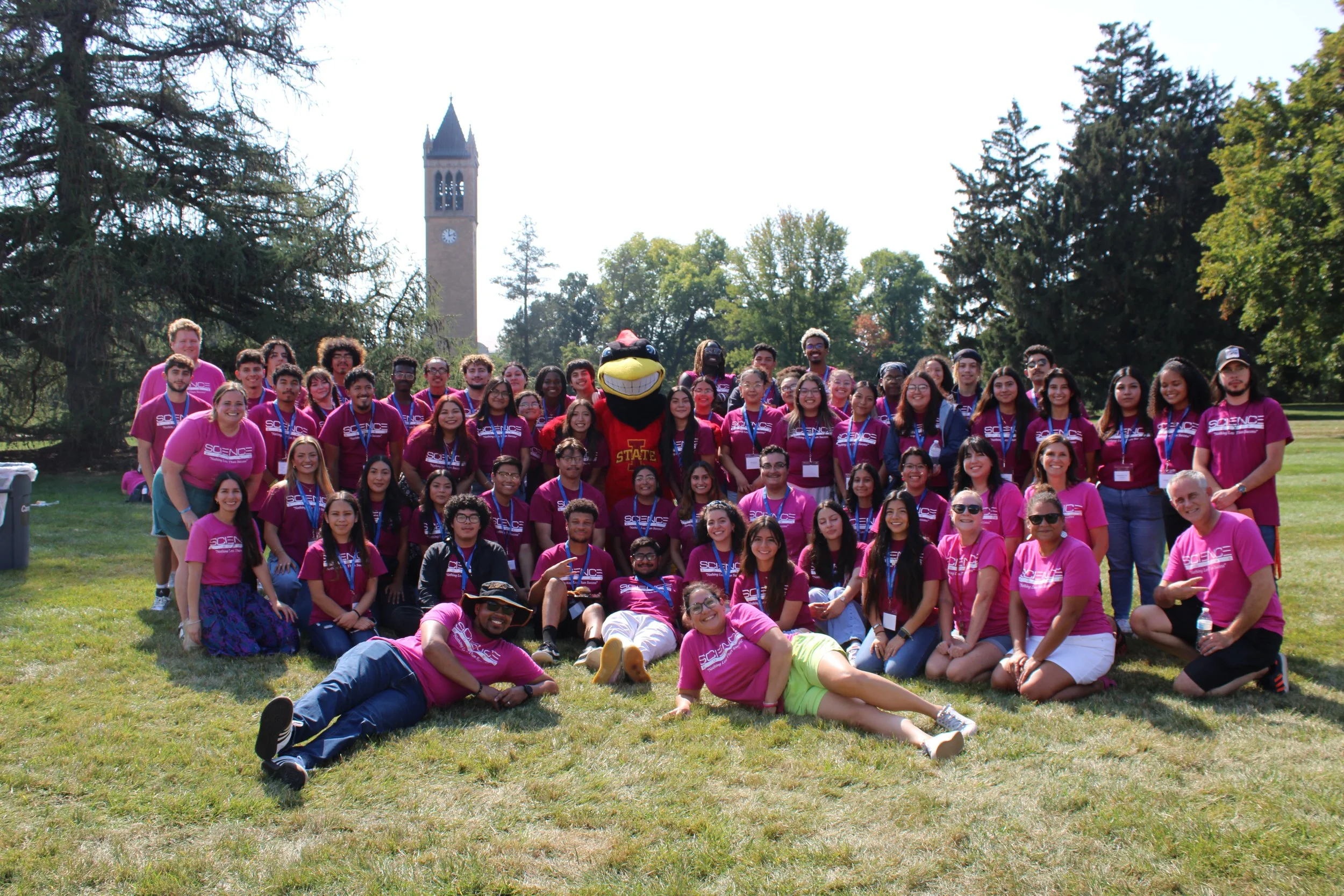 Group of people in red shirts posing outdoors with a mascot and a campanile in the background.