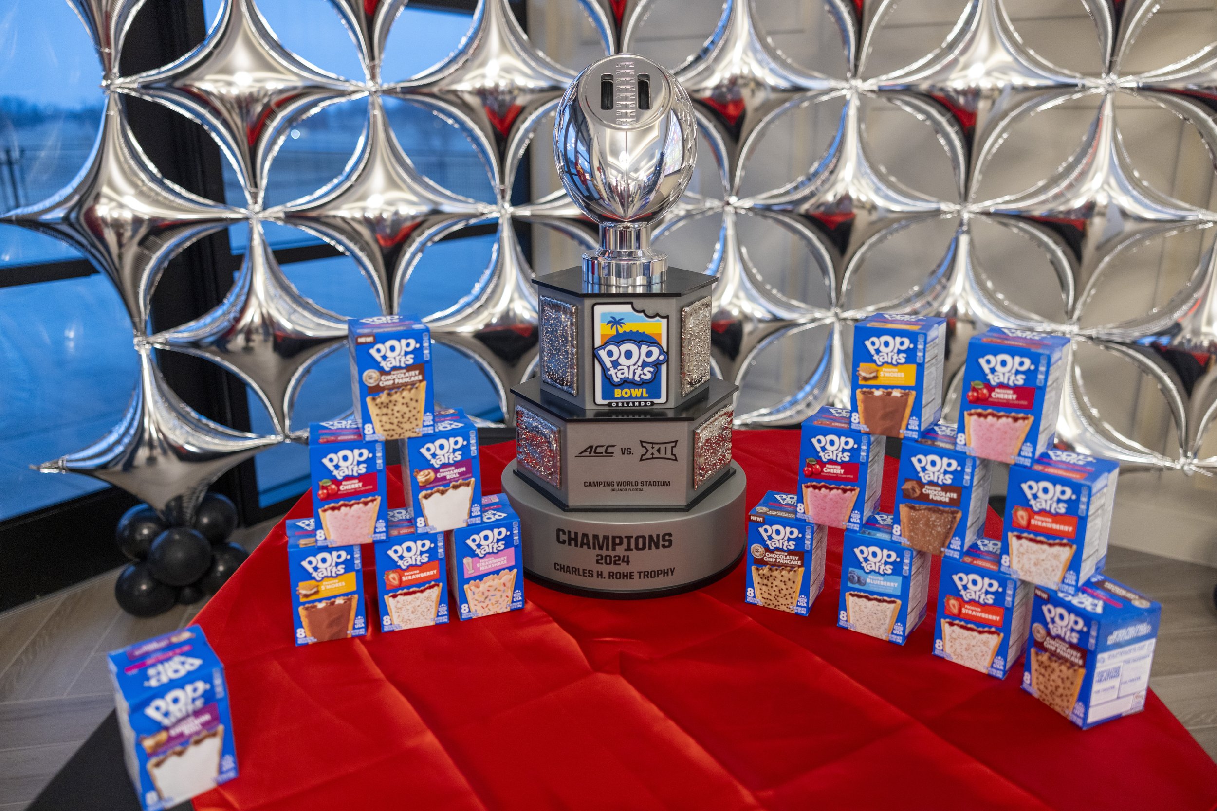 A display of Pop-Tarts boxes surrounding a trophy labeled "Pop-Tarts Bowl 2024 Champions Charles H. Rohe Trophy" on a red tablecloth with a silver balloon backdrop.