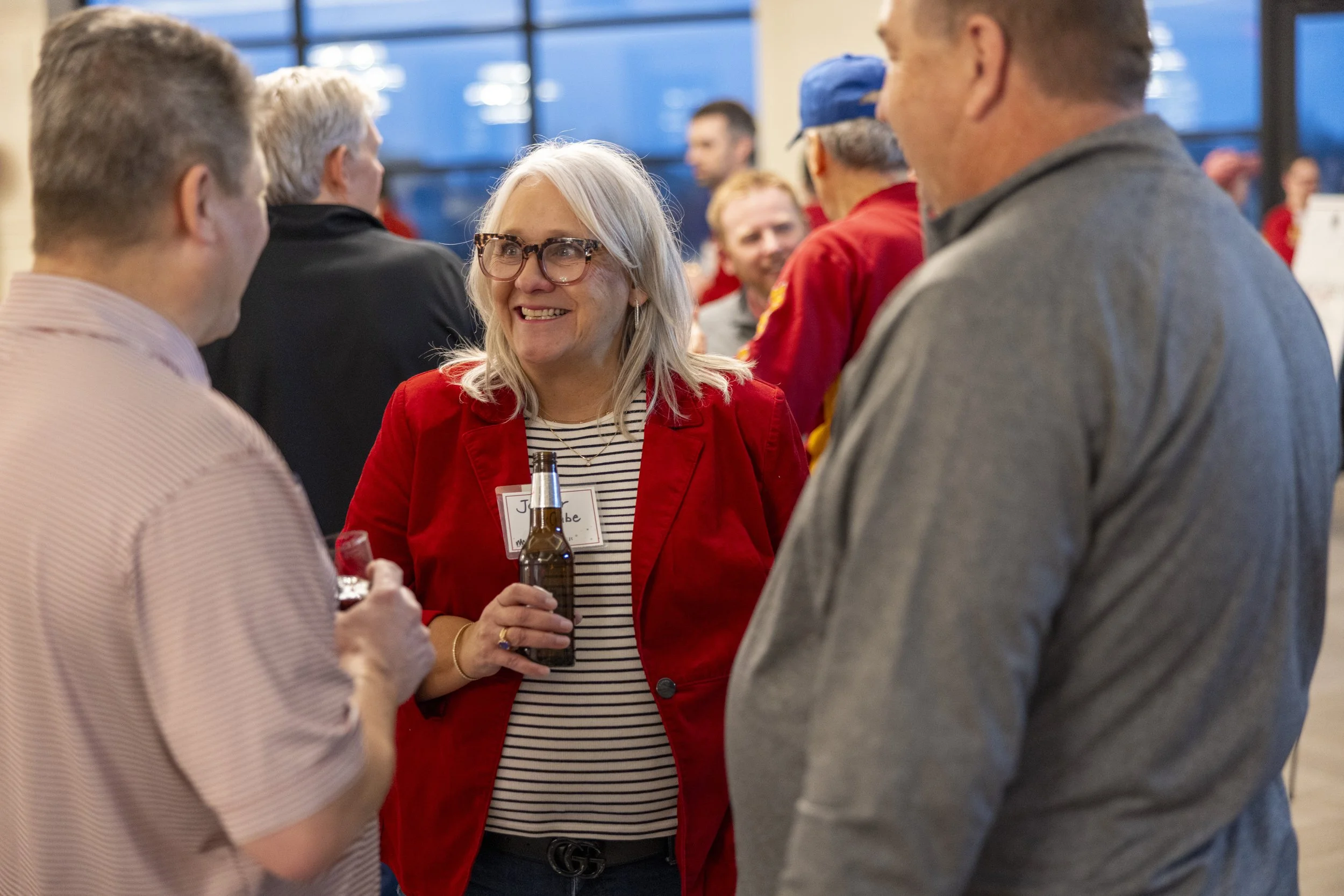 People socializing at a gathering, a woman in a red blazer holding a drink, smiling and talking to a man in a striped shirt.