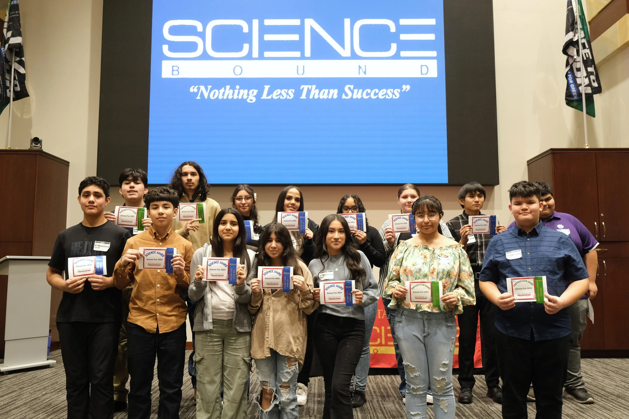 Group of students holding certificates at a Science Bound event with a "Nothing Less Than Success" banner in the background.