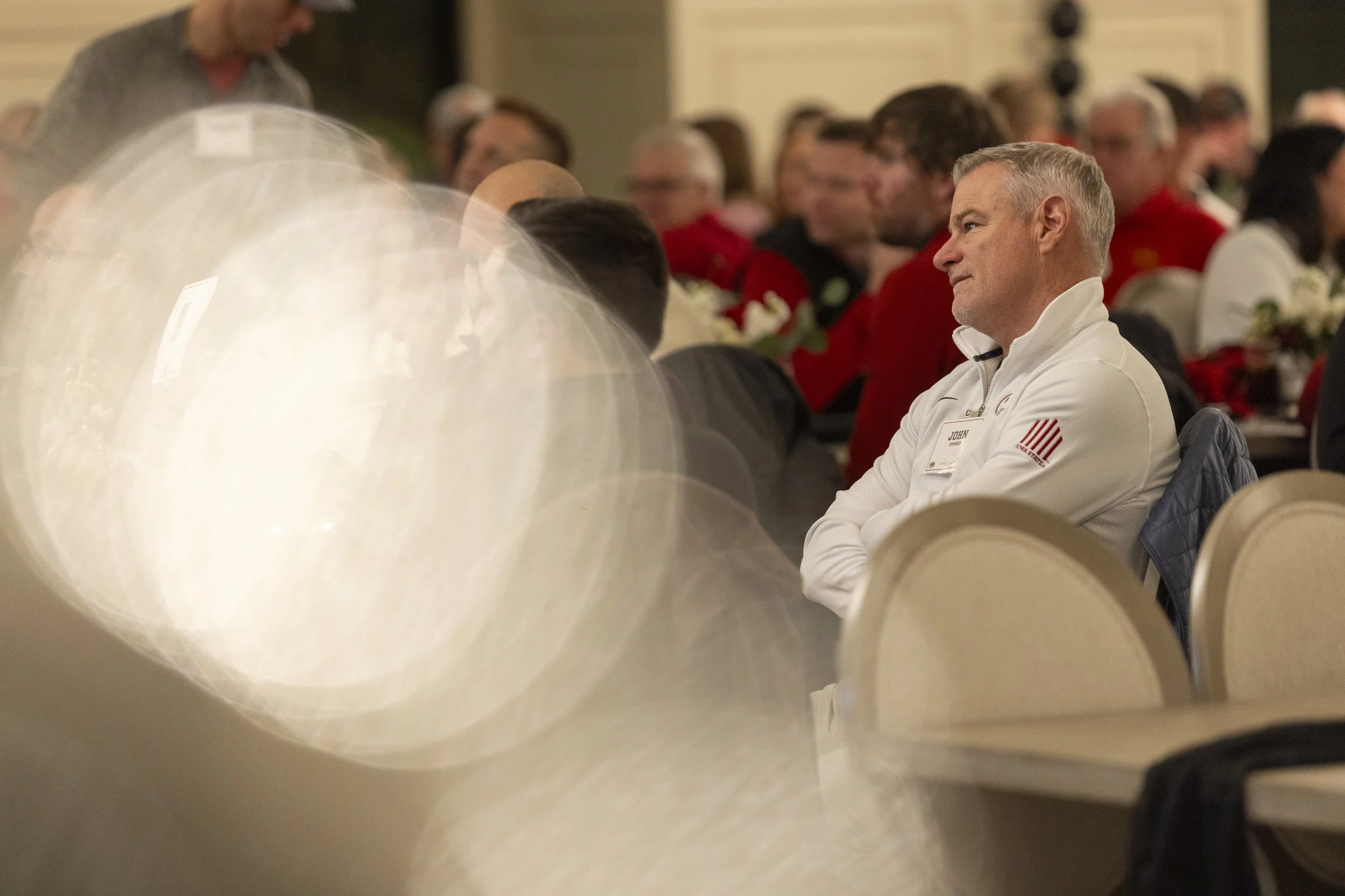 Man in white jacket seated at an indoor event with blurry foreground.