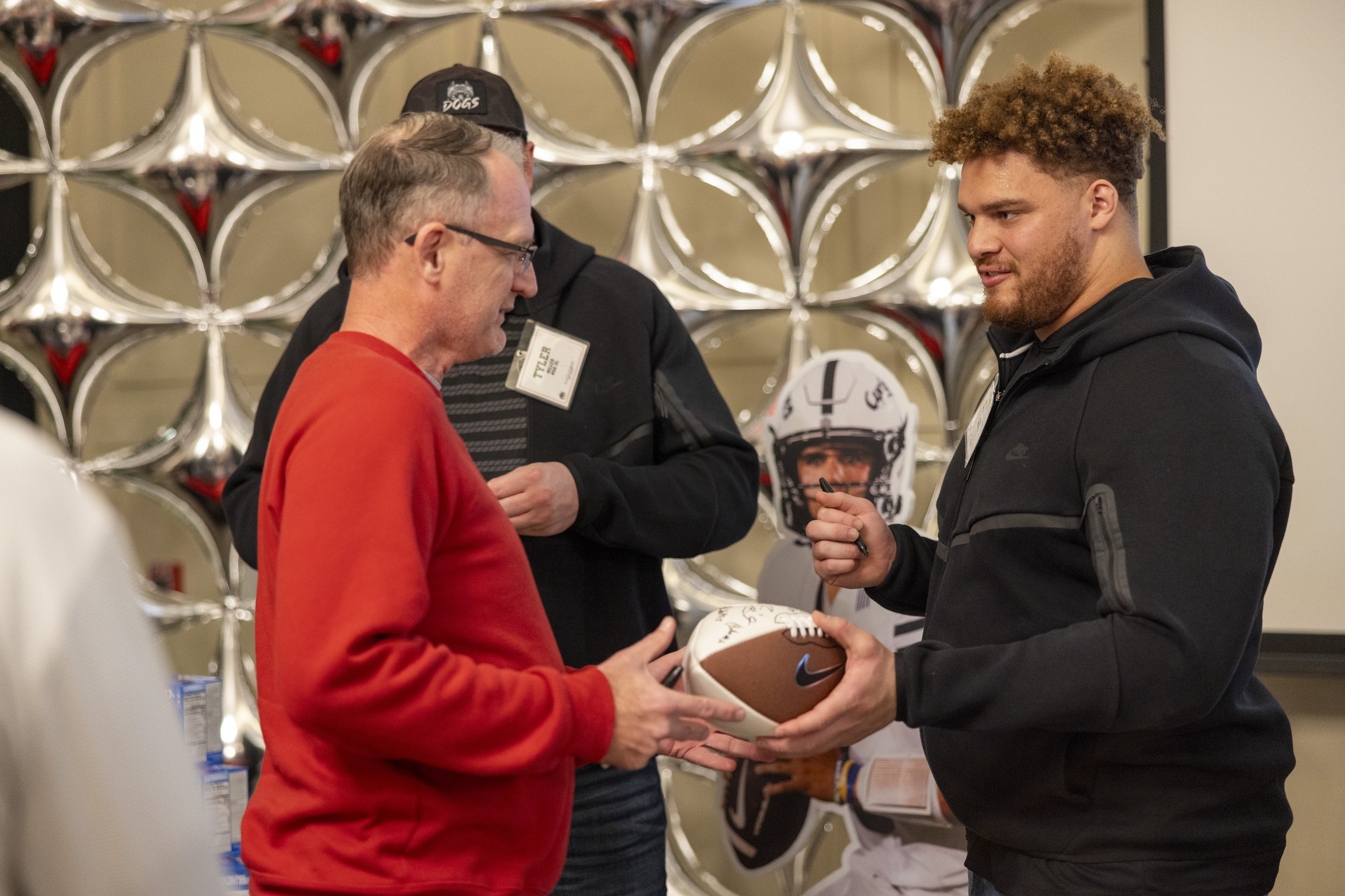 Two men exchanging a signed football indoors, with one man wearing a red sweatshirt and the other in a black hoodie. A cardboard cutout of a football player is visible in the background, set against a patterned wall.