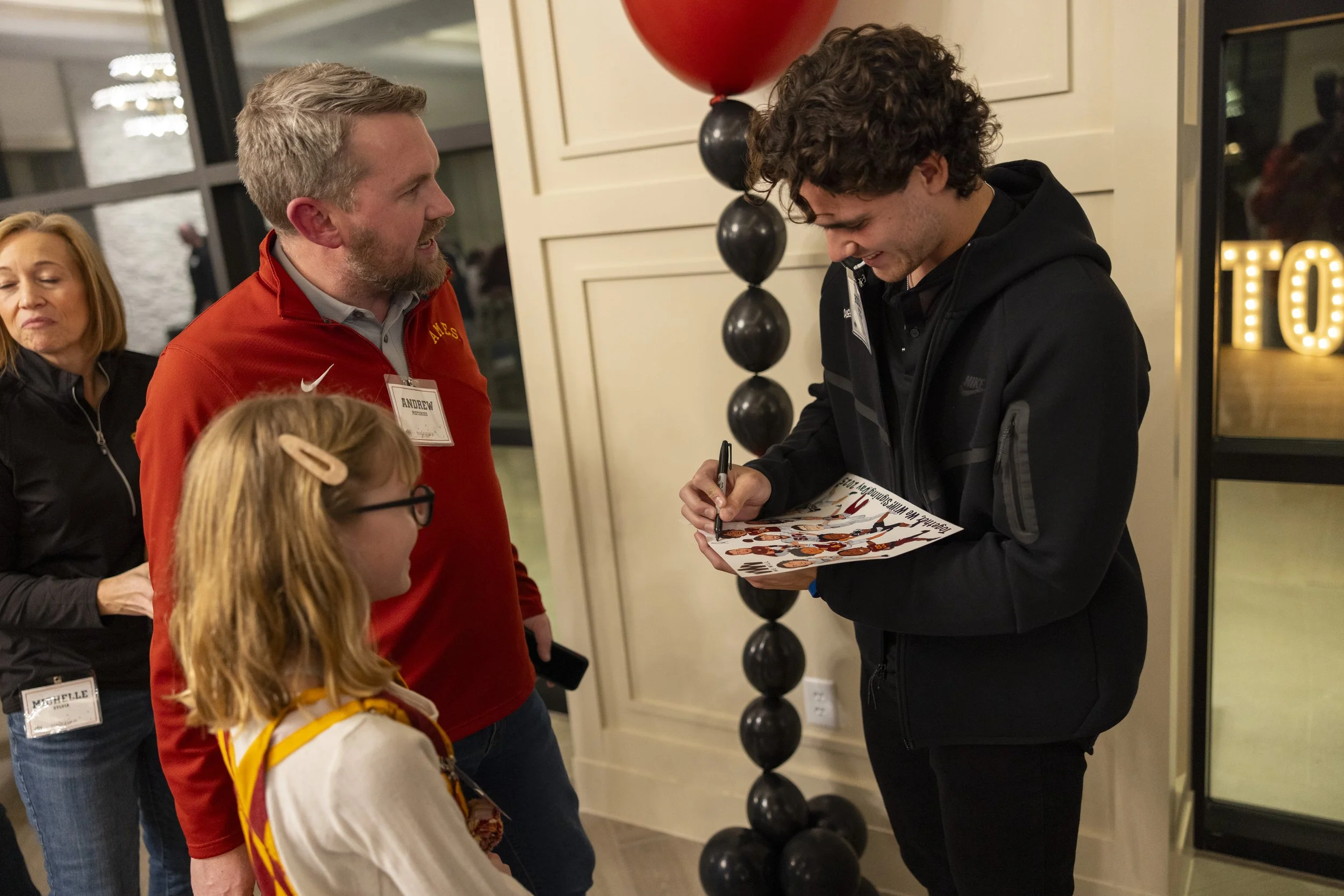 A person signing an autograph for a child, accompanied by two adults, with balloons and decorations in the background.