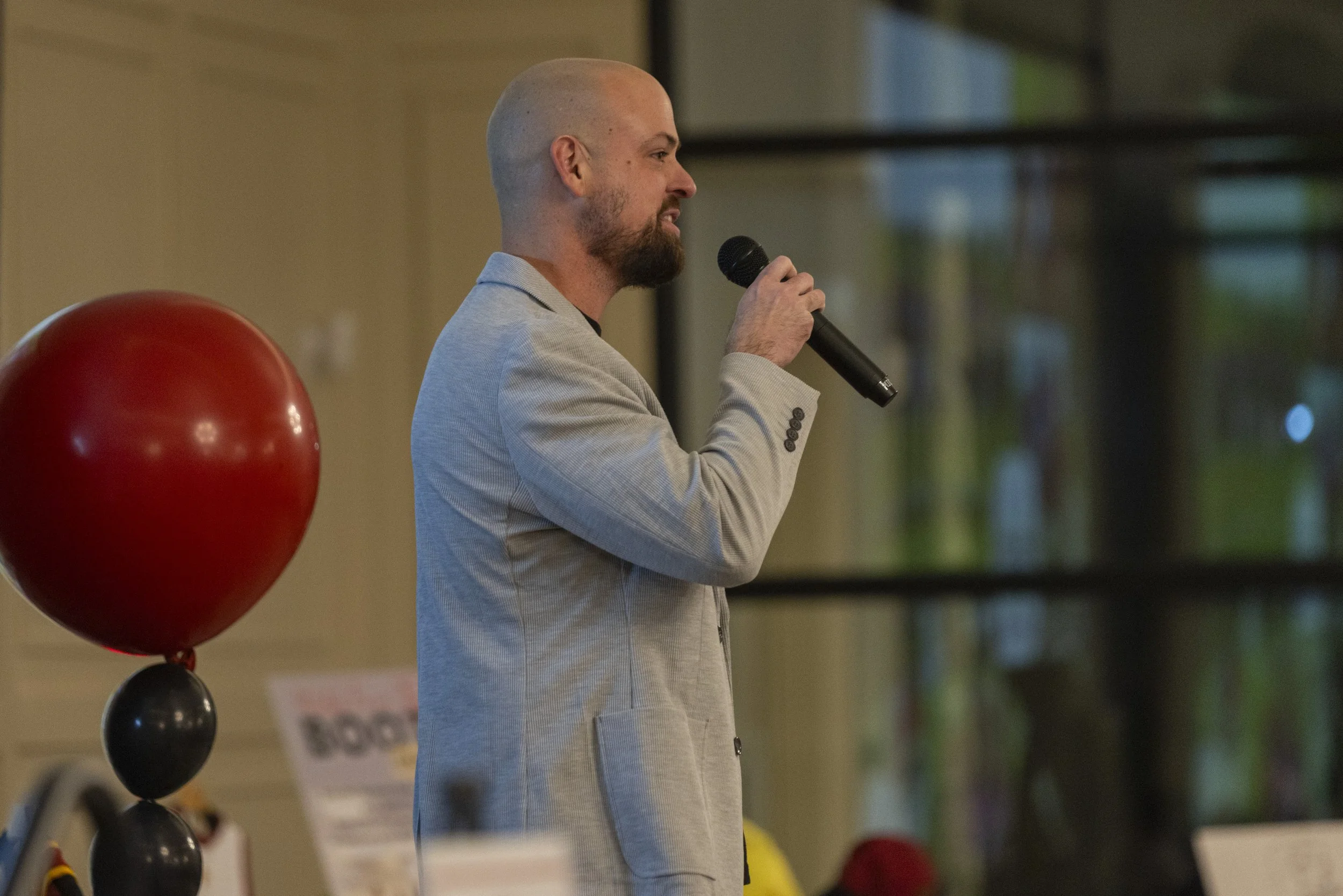 Man in a light gray suit speaking into a microphone indoors, with a red and black balloon decoration nearby.