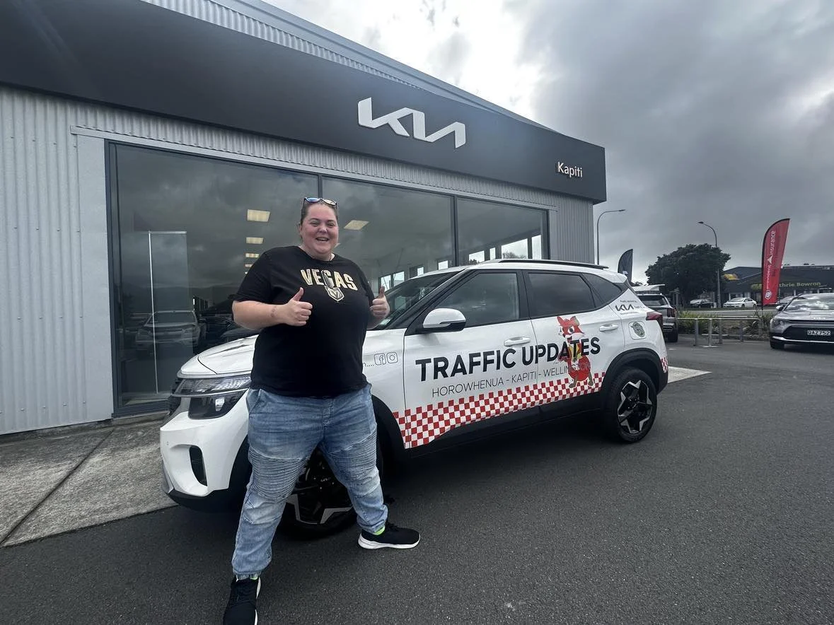 A person giving thumbs up in front of a white car with "Traffic Updates" signage parked outside a Kia dealership.