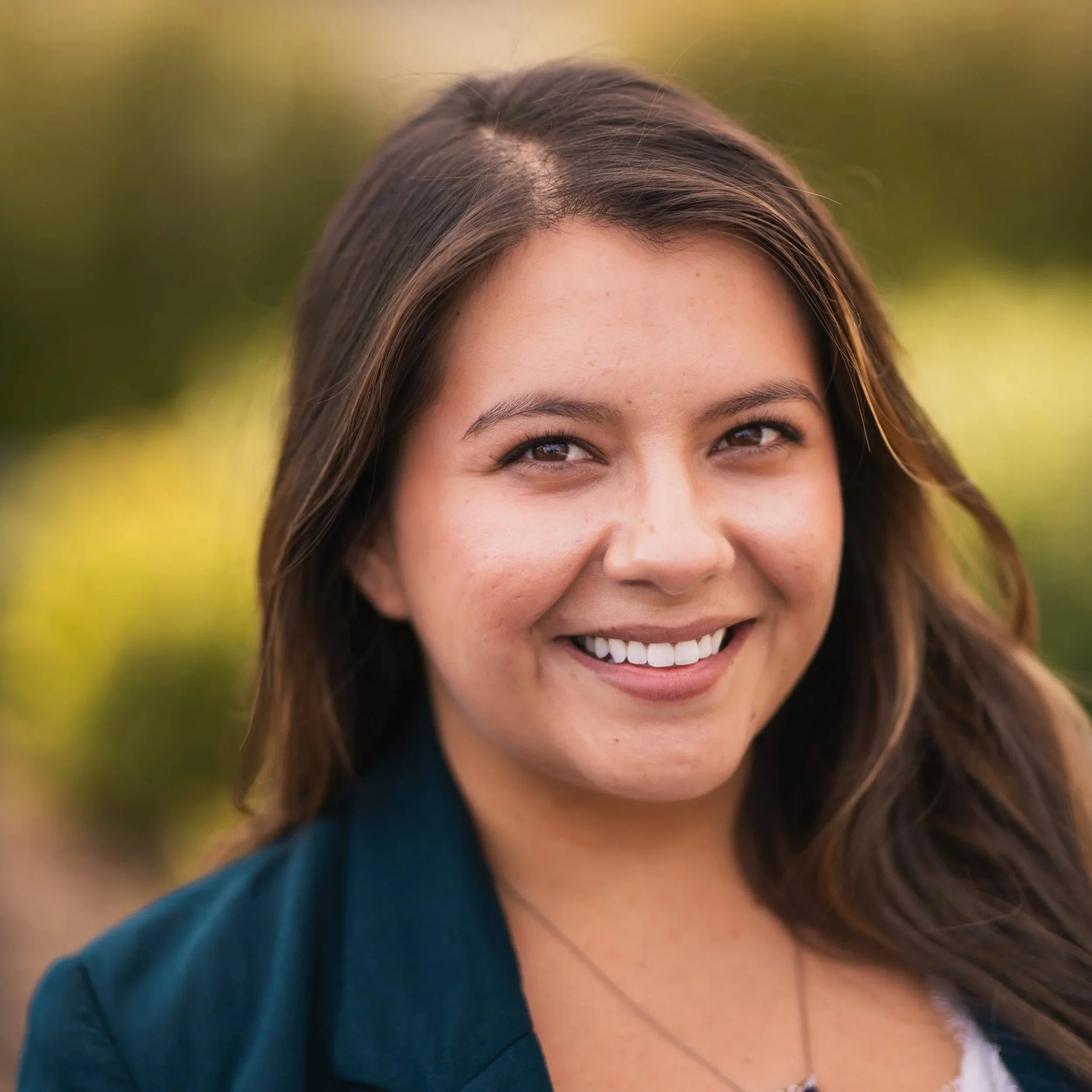 Close-up of a smiling woman outdoors with blurred green foliage in the background.