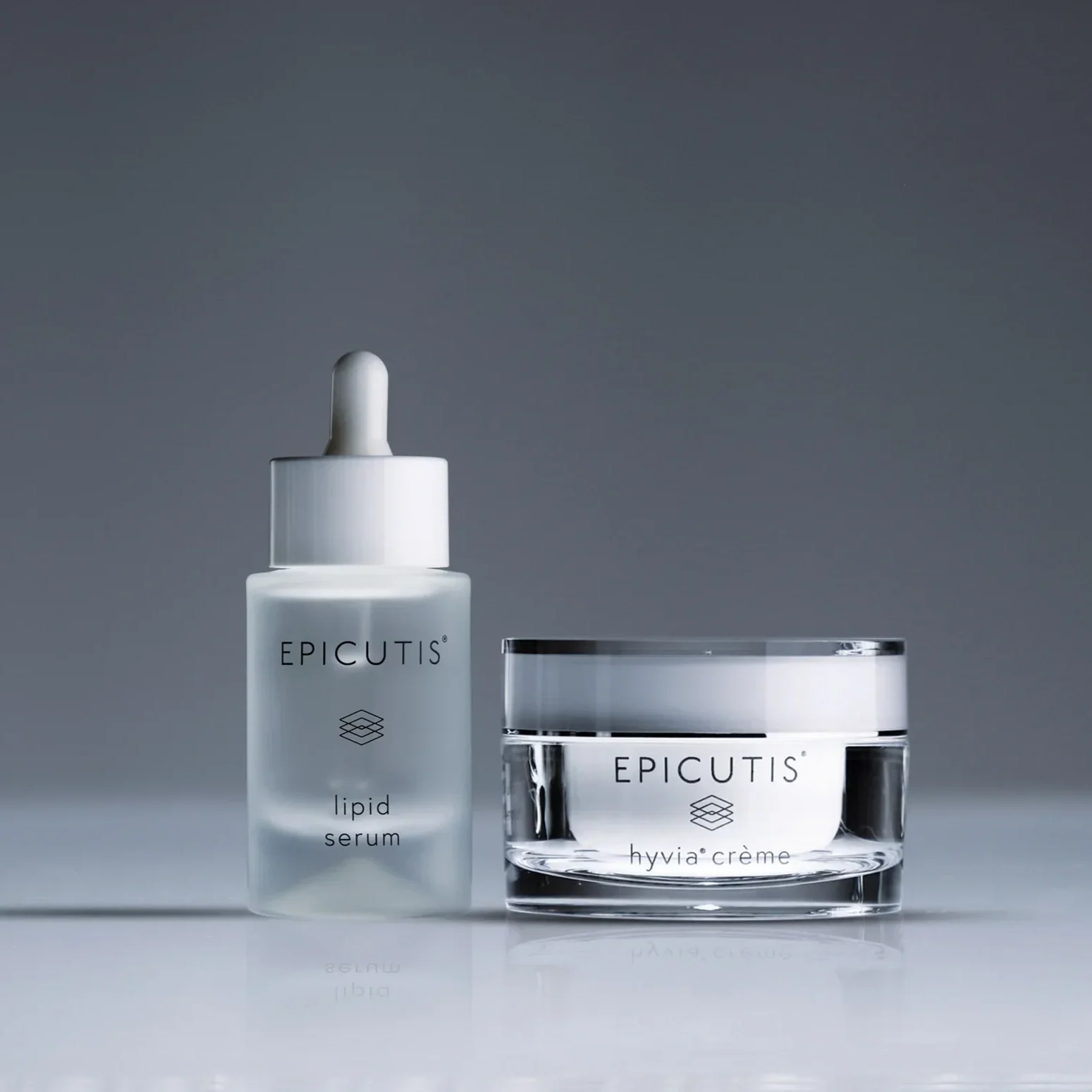 Two skincare products: a frosted glass bottle with a dropper labeled 'Epicutis lipid serum' and a clear jar labeled 'Epicutis hyvia crème' on a reflective surface against a gray background.