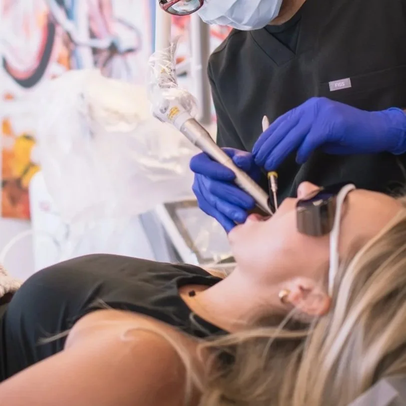 A woman lying down and receiving a laser therapy from a dentist wearing gloves and a face mask .