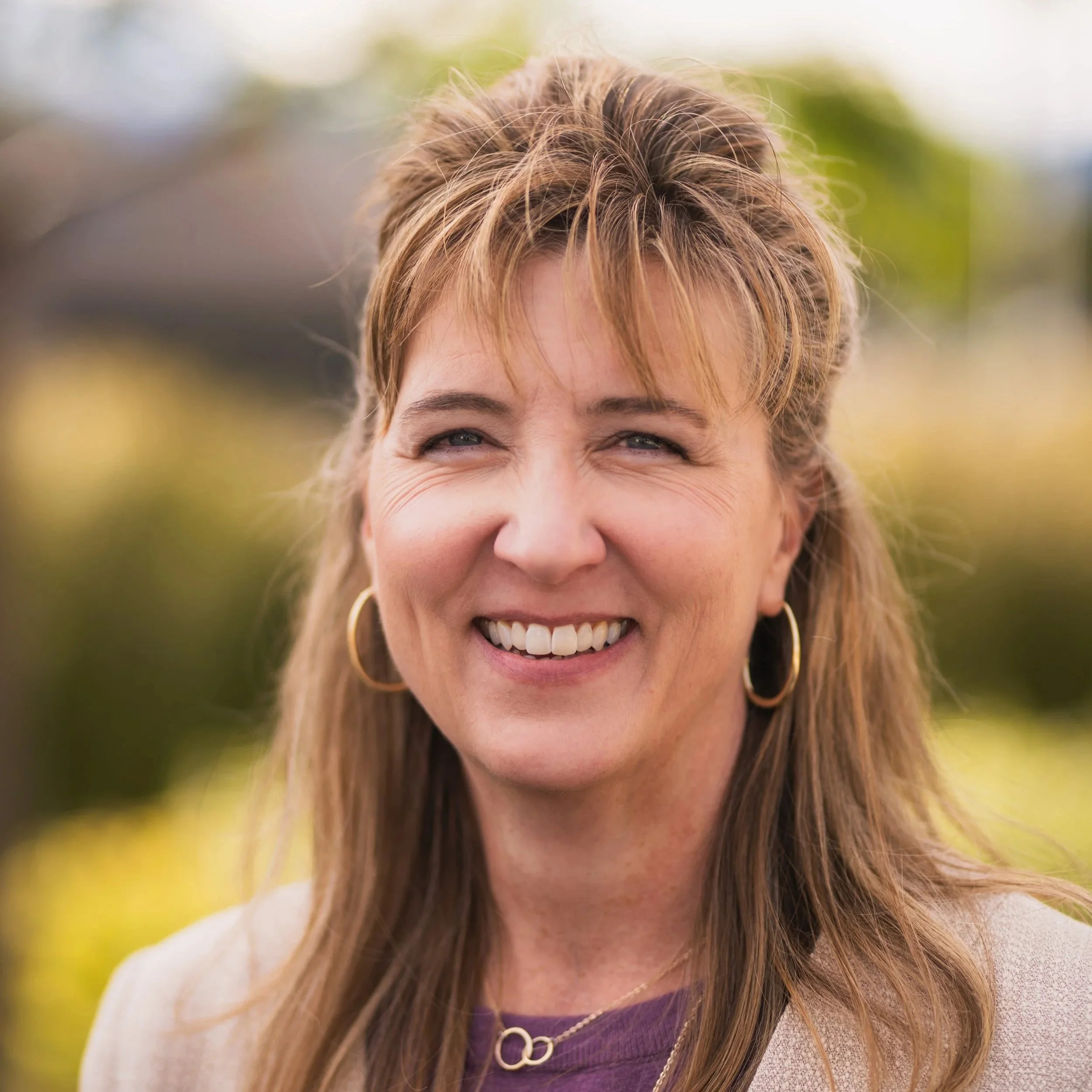 Smiling woman with light brown hair and gold hoop earrings outdoors.