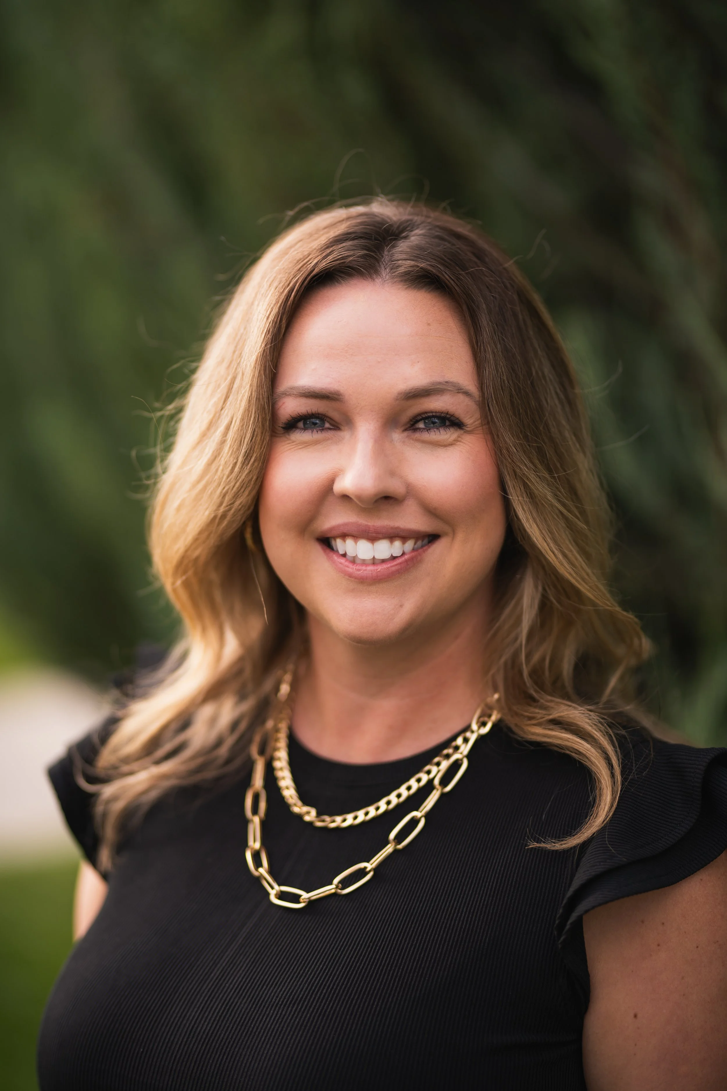 A smiling woman with shoulder-length wavy brown hair, wearing a black top with ruffled sleeves and gold layered chain necklaces, standing outdoors with blurred green foliage in the background.