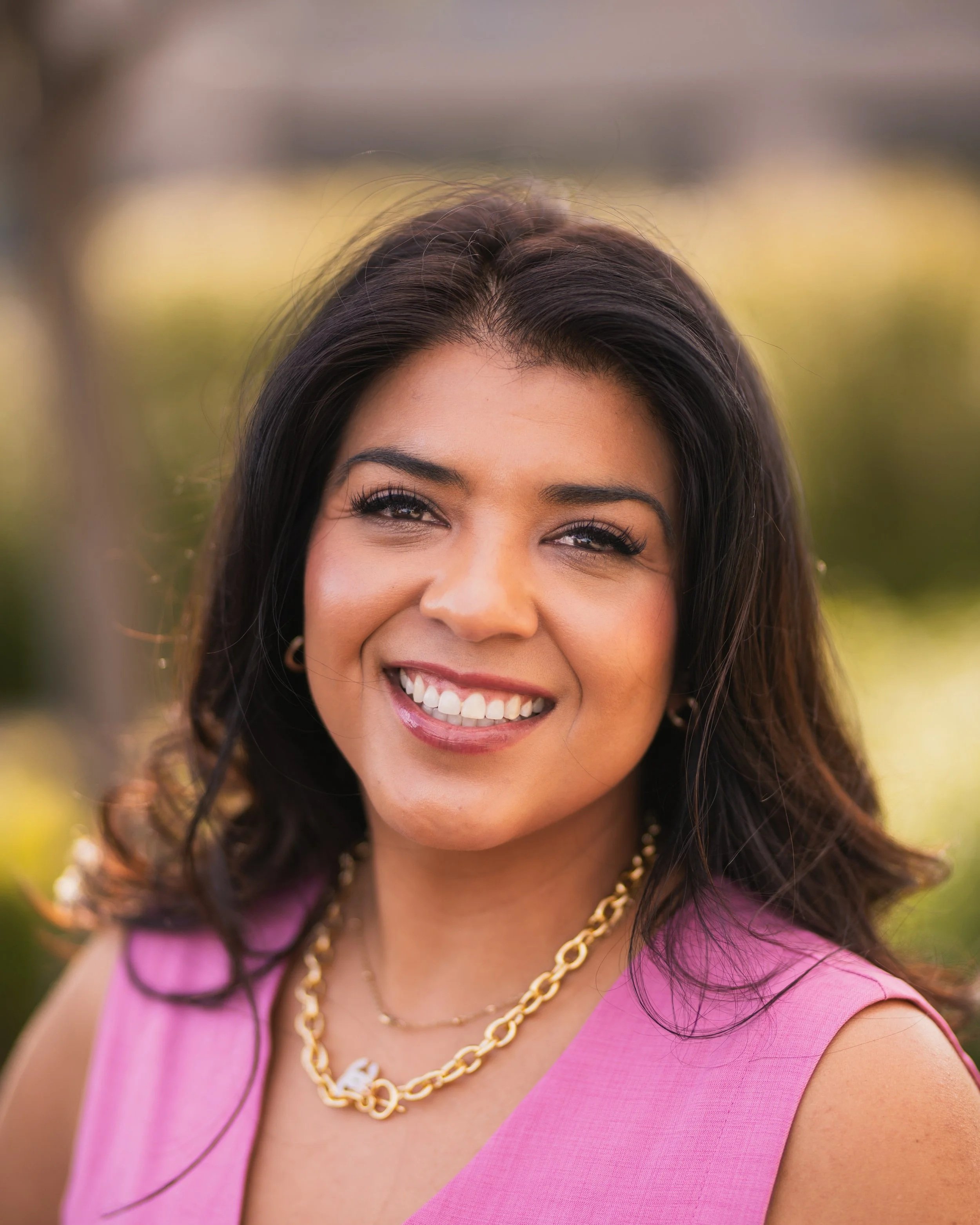A woman with dark hair and a bright smile, wearing a pink sleeveless top, gold chain necklace, and earrings, outdoors with blurred greenery in the background.