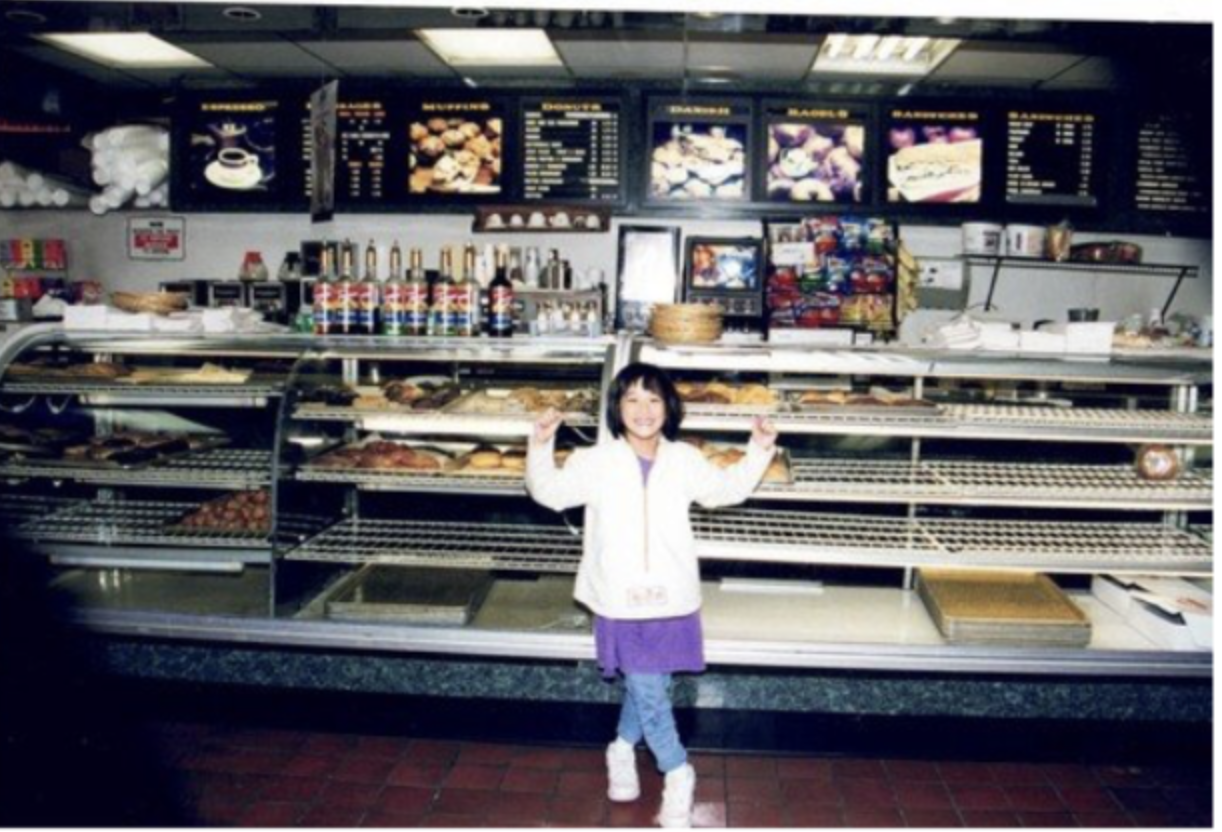 A young Dorothy Chow smiling and posing in front of the donut display case in her parents donut shop.