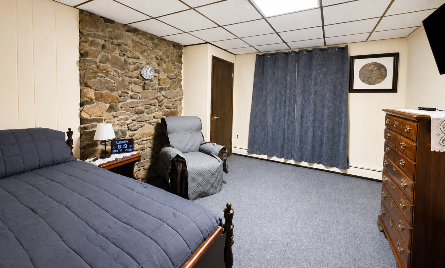 A bedroom with a stone accent wall, blue curtains, a wooden dresser, a bed with a blue comforter, a gray recliner, a nightstand with a lamp and digital clock, and framed art on the wall.