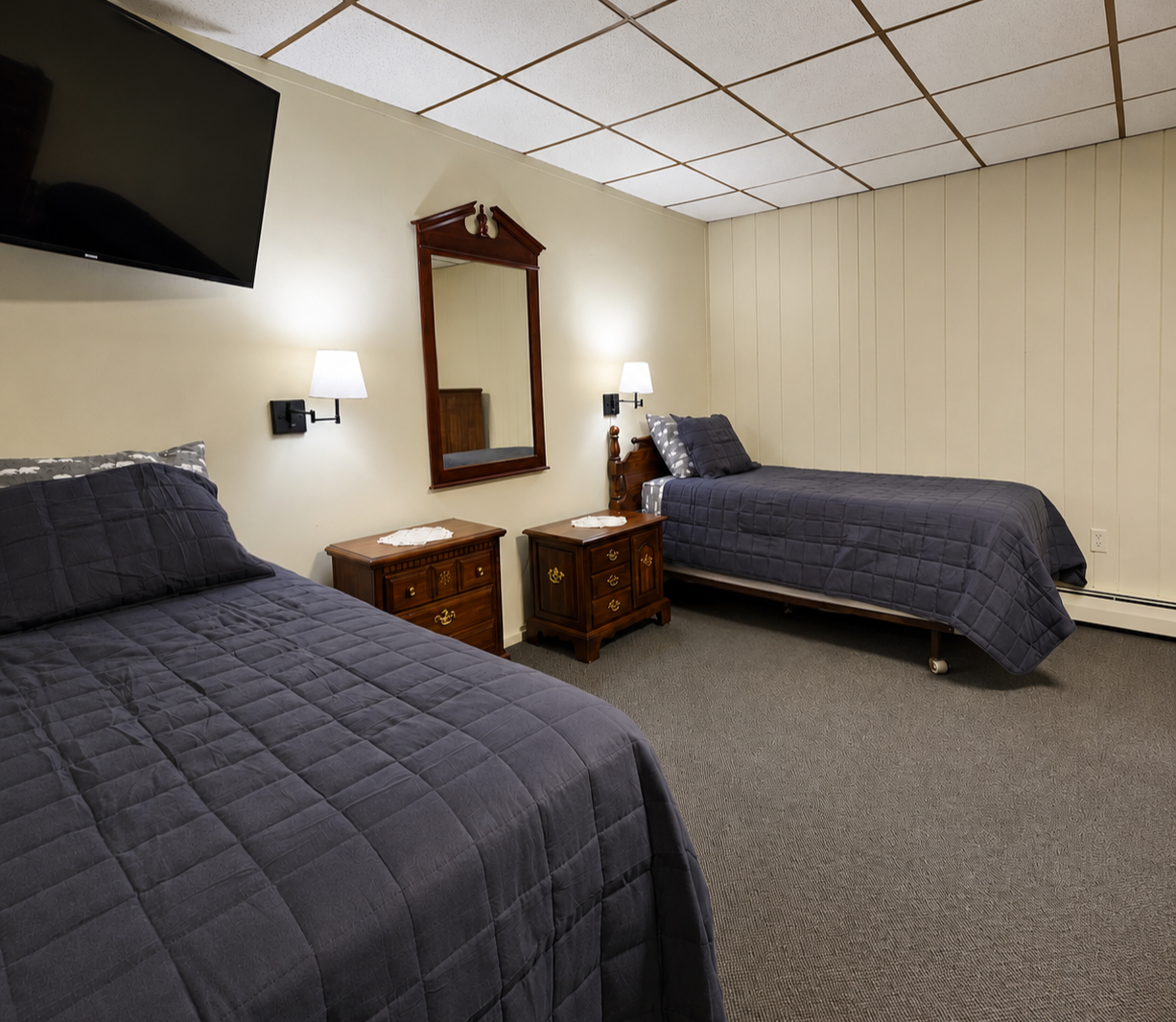 Room with two twin beds with dark gray quilts, two wooden nightstands with lace doilies, a wall mirror, a wall-mounted lamp, and a flat-screen TV on the wall.