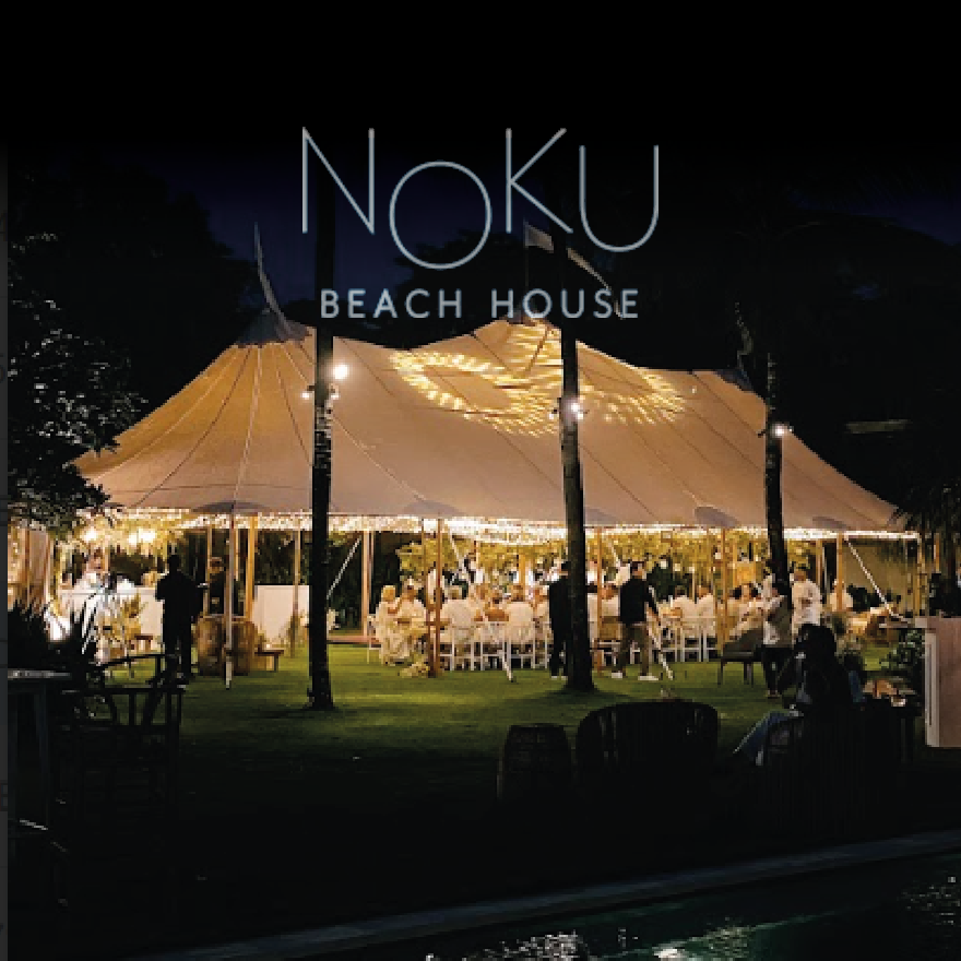 A night shot of a wedding dinner in a tent at Noku Beach House in Bali. The tent is decorated with white lights and flowers.The decorations are beautifully lighted up, creating a festive and romantic atmosphere.