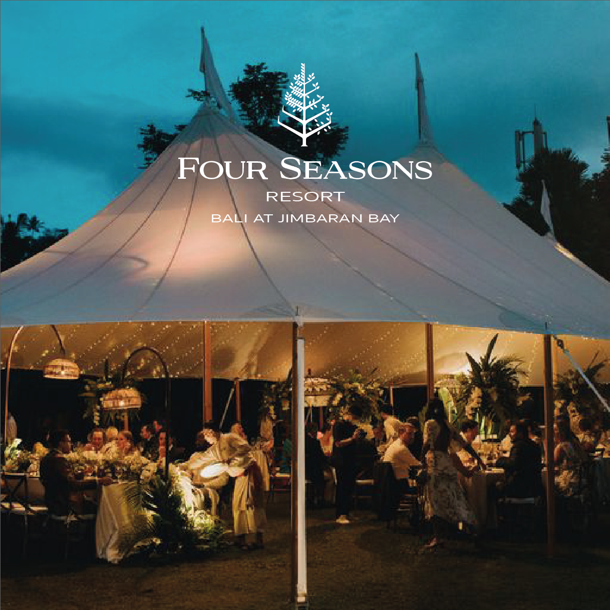 A night shot of a wedding dinner in a tent at the Four Seasons Resort Bali at Jimbaran Bay. The tent is decorated with white lights and flowers. The guests are dressed in formal attire and are enjoying a delicious meal.