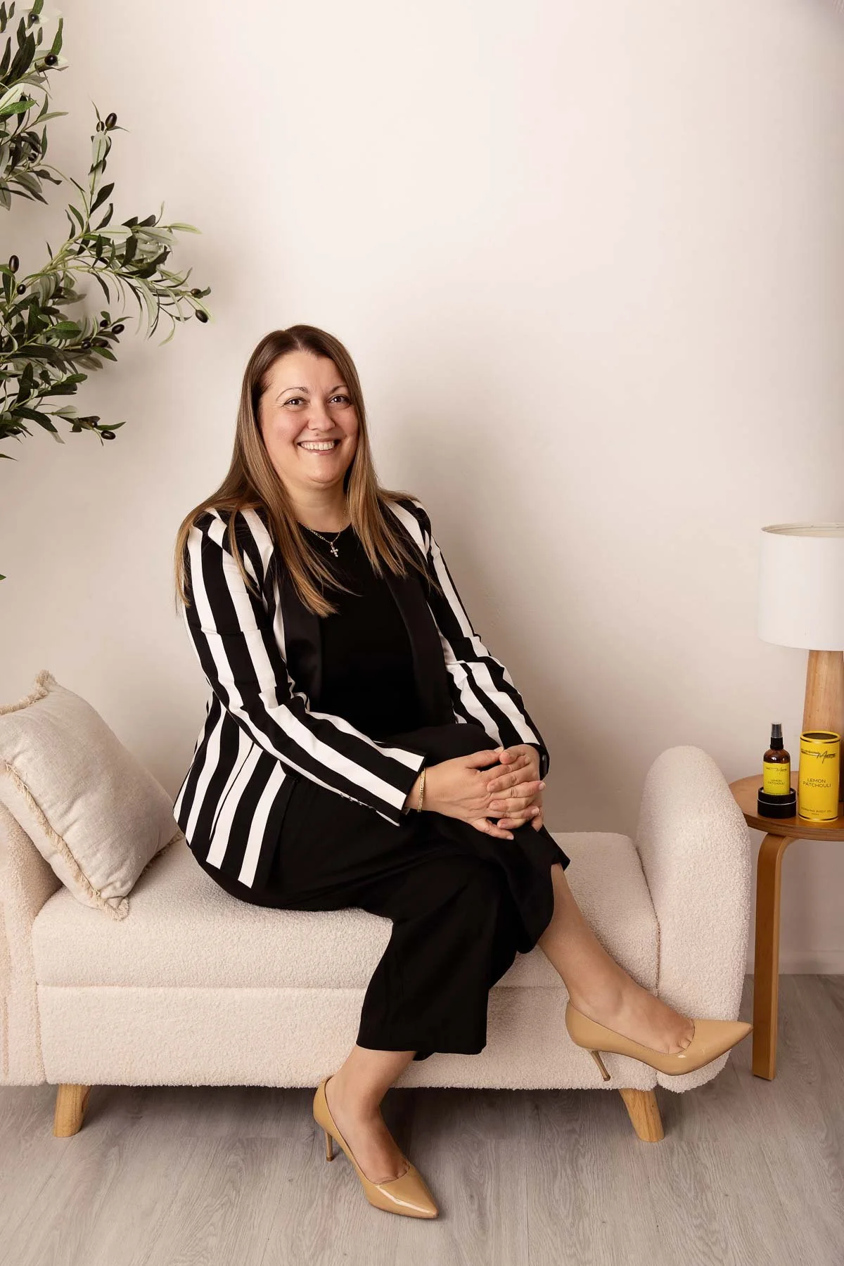 A woman with blonde hair wearing a black & white jacket, sitting cross-legged on a beige sofa, smiling at the camera in a cozy, well-lit room.