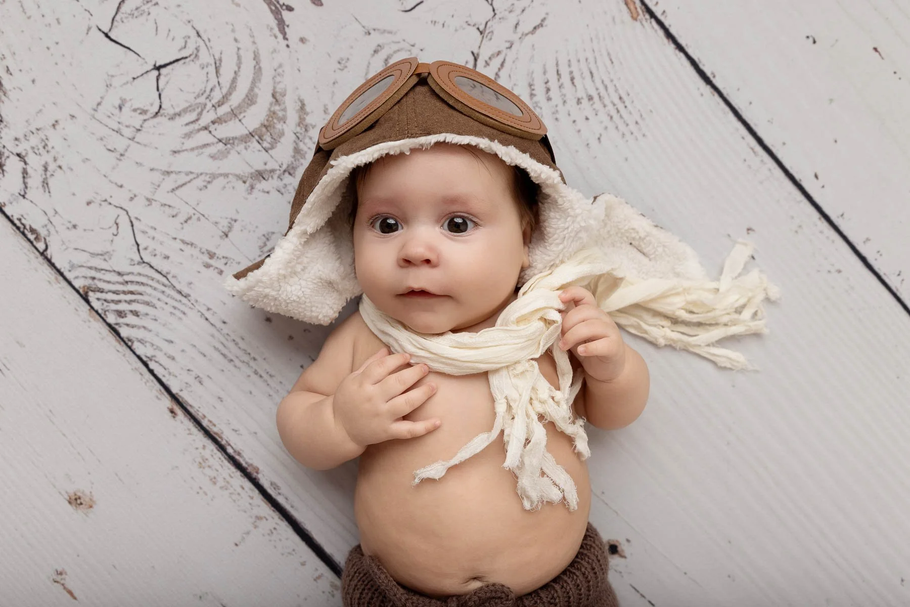 Baby wearing aviator hat and scarf, lying on wooden floor
