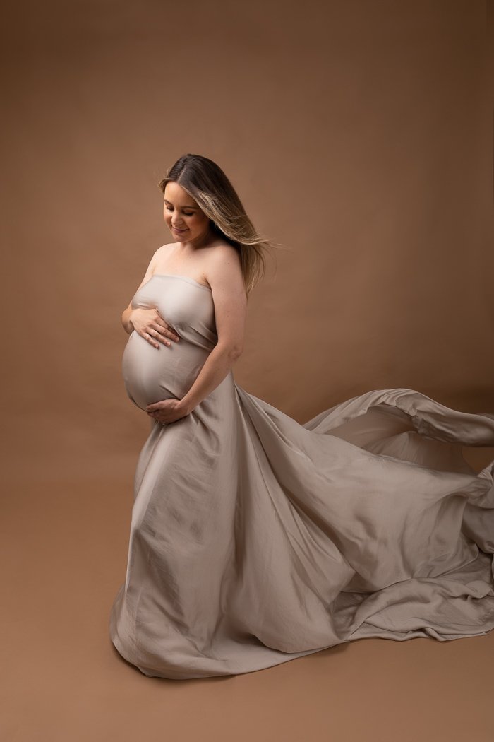 Pregnant woman in flowing gown holding belly against a brown background.