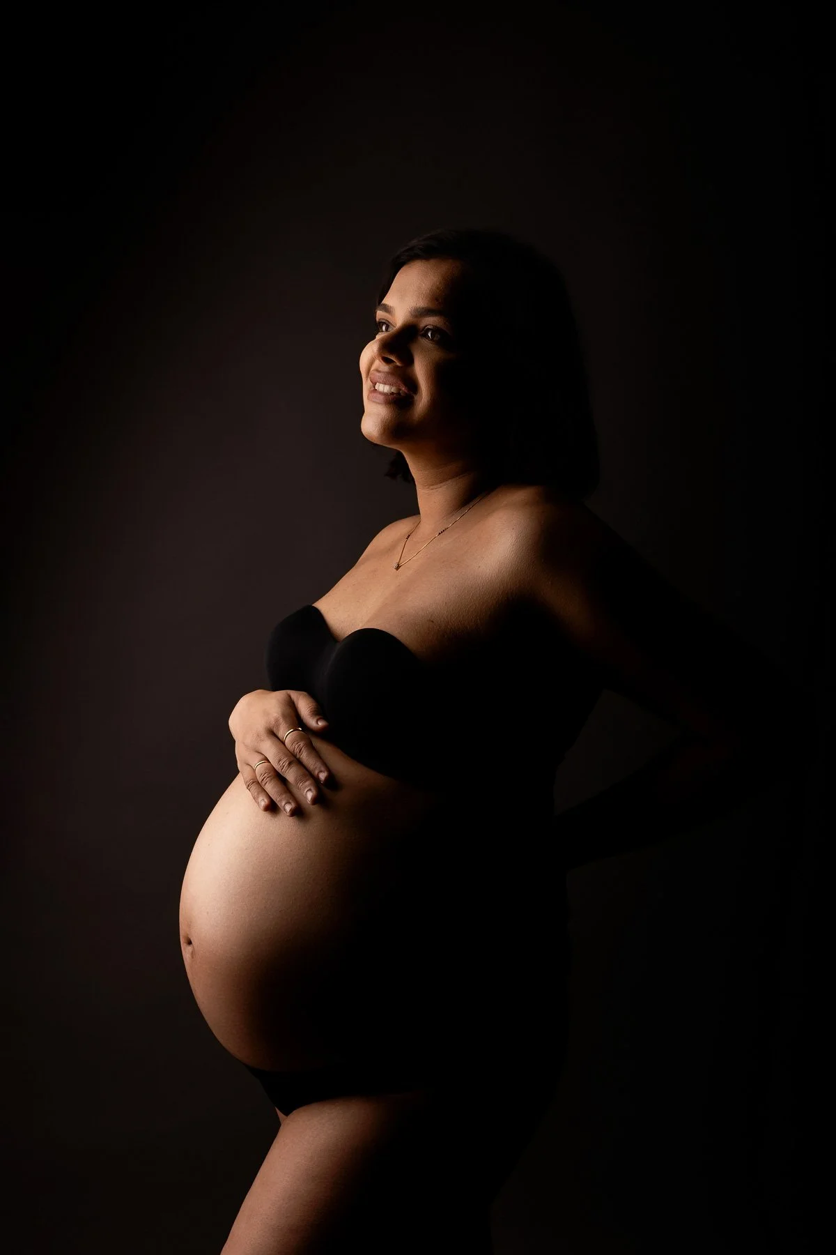 Pregnant woman in black maternity wear, holding her belly against a dark background. maternity photographer North Shore Sydney.