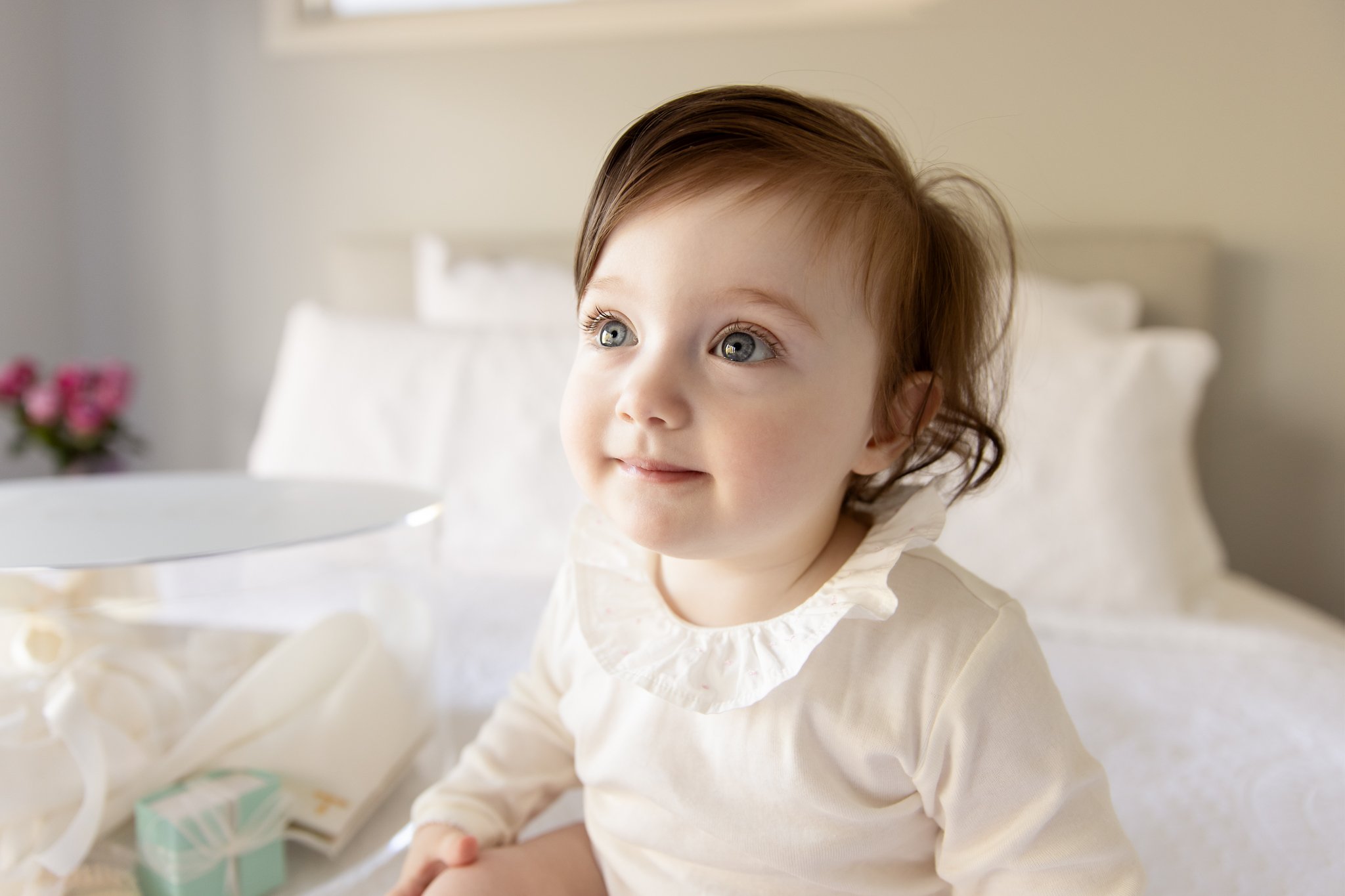 Smiling baby wearing white outfit with ruffled collar on bed.