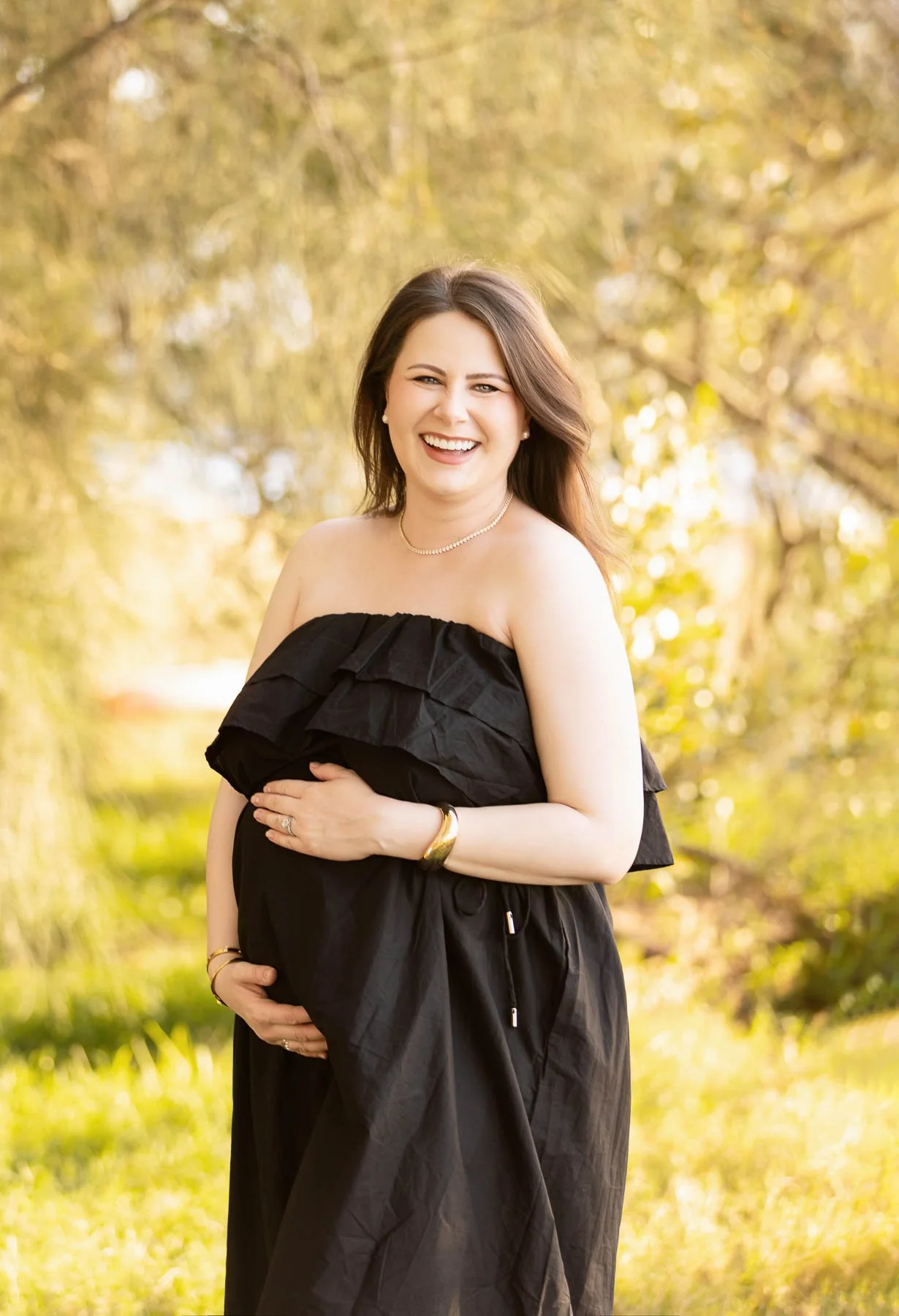 Pregnant woman smiling wearing a black dress with trees behind her