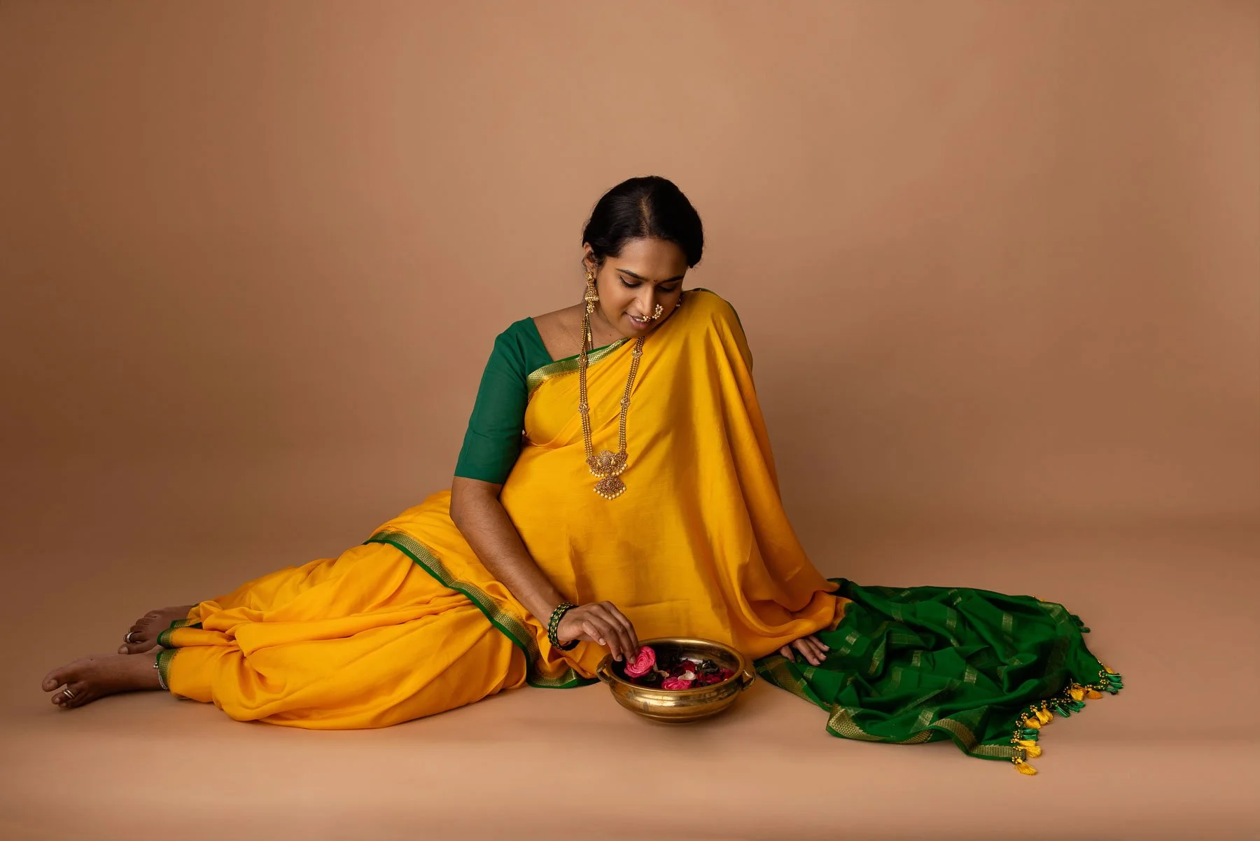 Pregnant woman in yellow & green saree, wearing golden jewelries, holding her belly, against a warm neutral background.