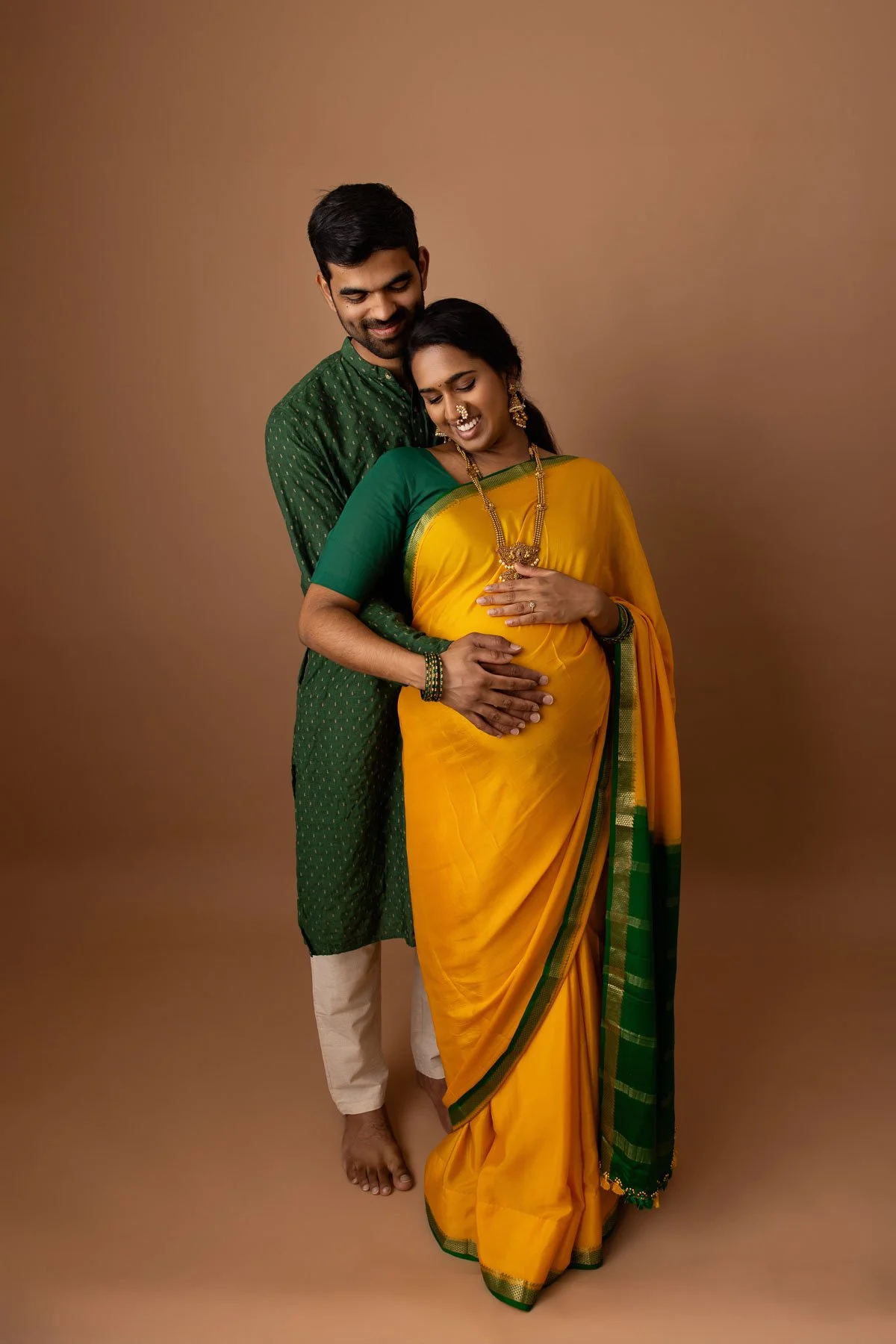 Pregnant woman in yellow & green saree, wearing golden jewelries, holding her belly, against a warm neutral background.