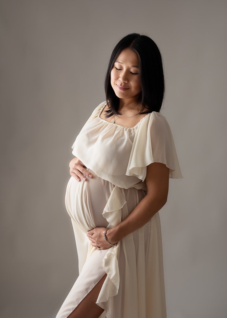 Pregnant woman in cream dress holding belly, looking down, with soft lighting and neutral background.