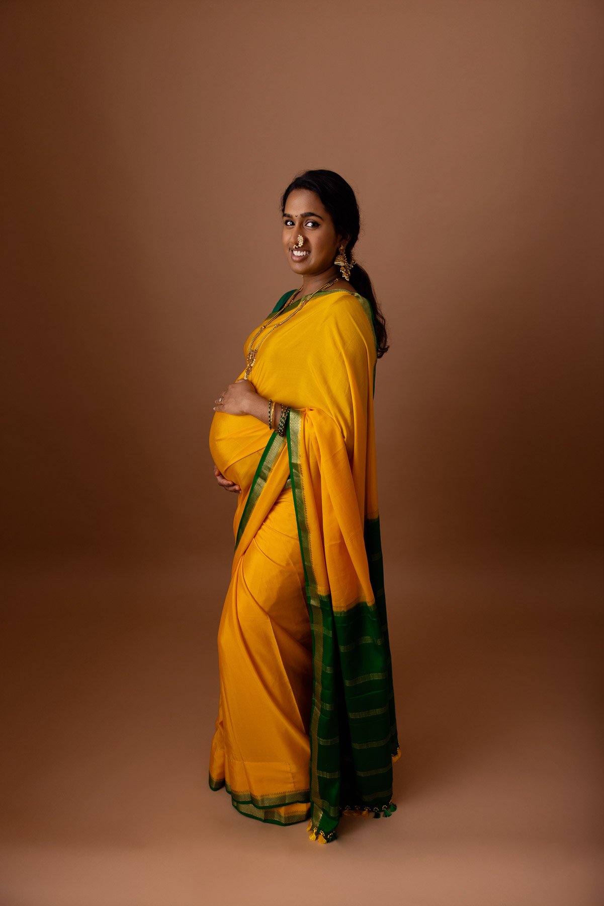 Pregnant woman in yellow & green saree, wearing golden jewelries, holding her belly, against a warm neutral background.